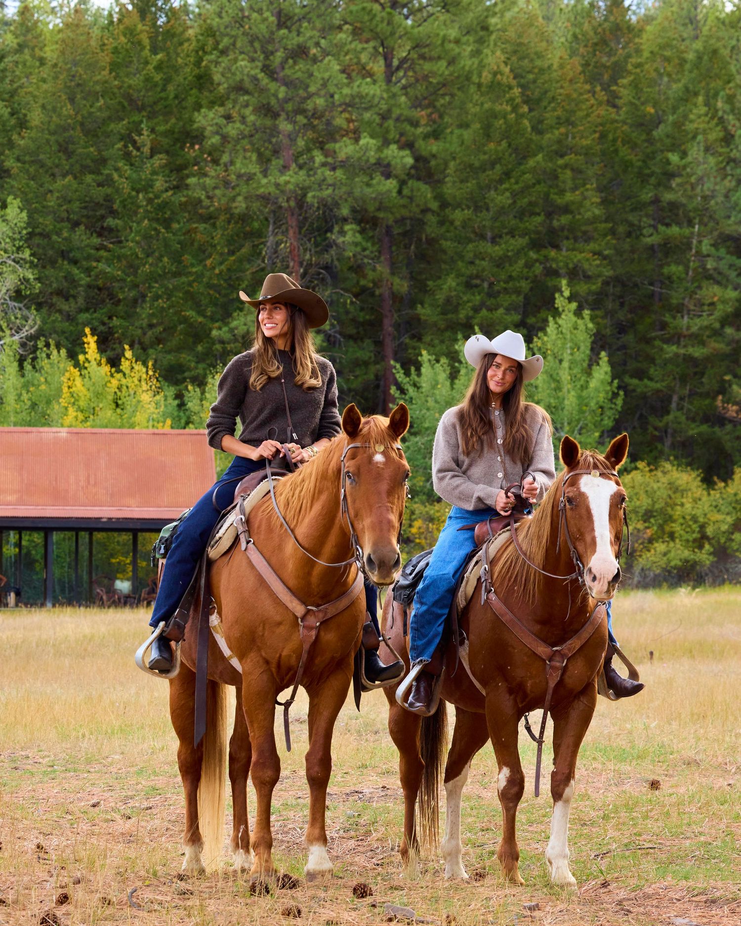 Two women in cowboy hats riding chestnut horses in open field with forest backdrop.