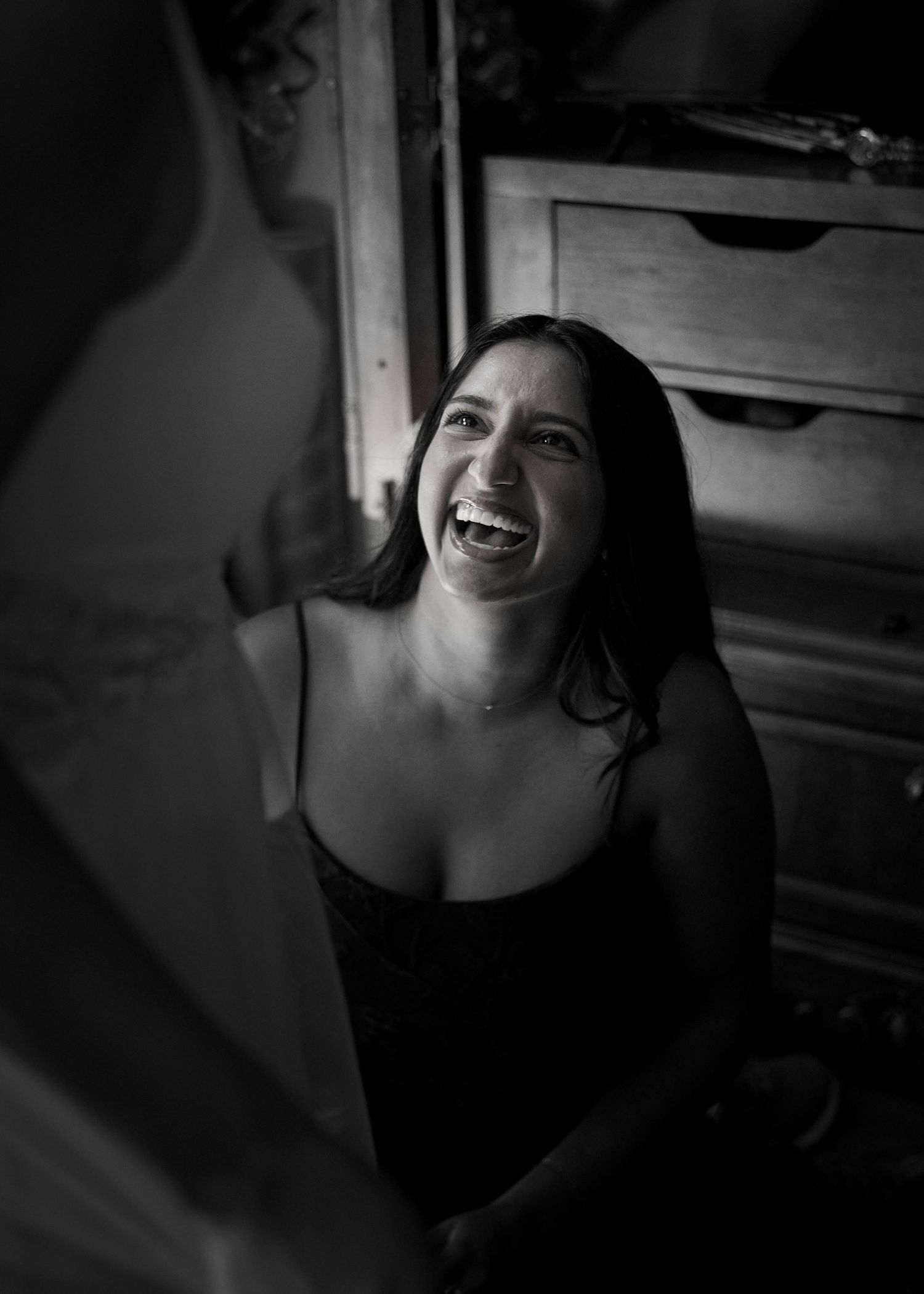 Black and white photo of a laughing woman with dark hair sitting near a wooden dresser.