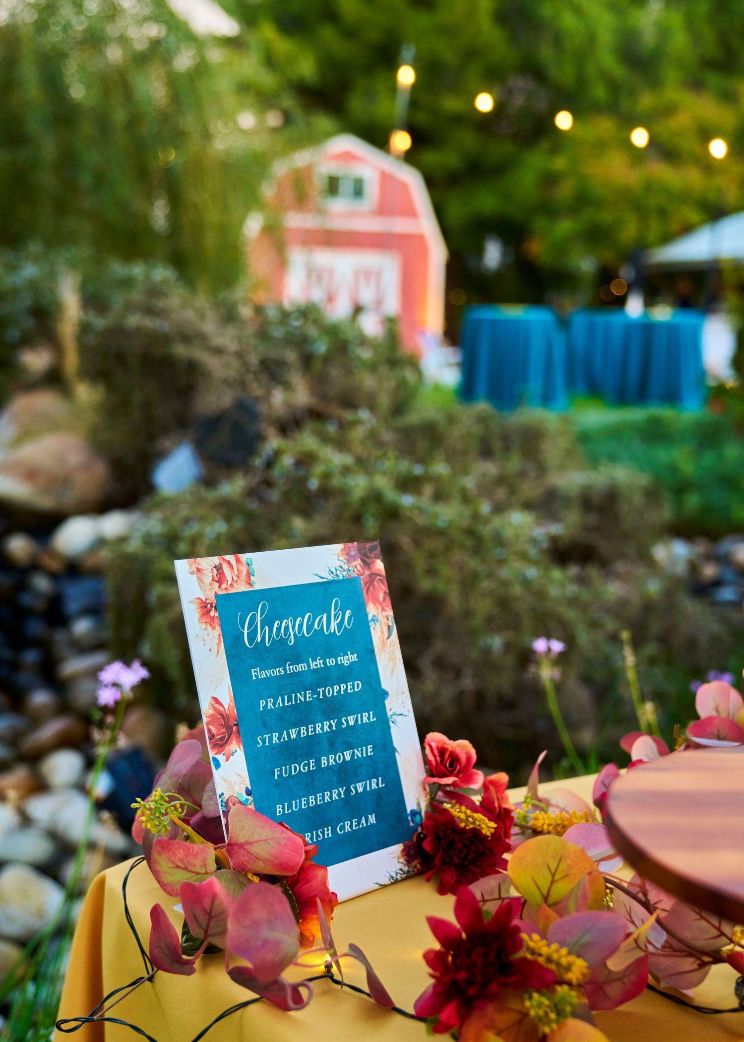 Rustic outdoor cheesecake dessert table sign with floral decor, string lights, and red barn backdrop at a fall wedding.