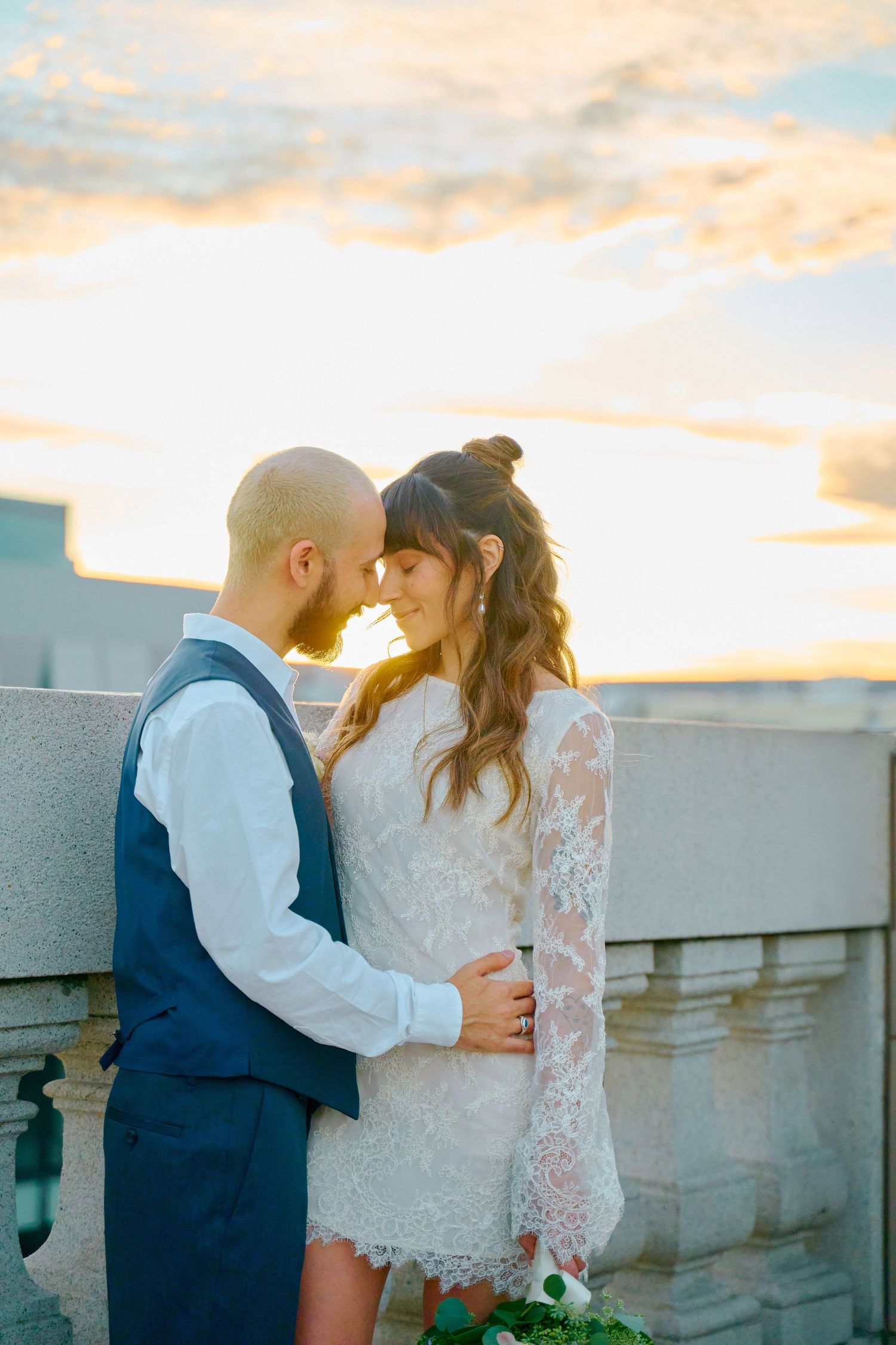 Couple embraces forehead-to-forehead in golden sunset light on a rooftop terrace in wedding attire.
