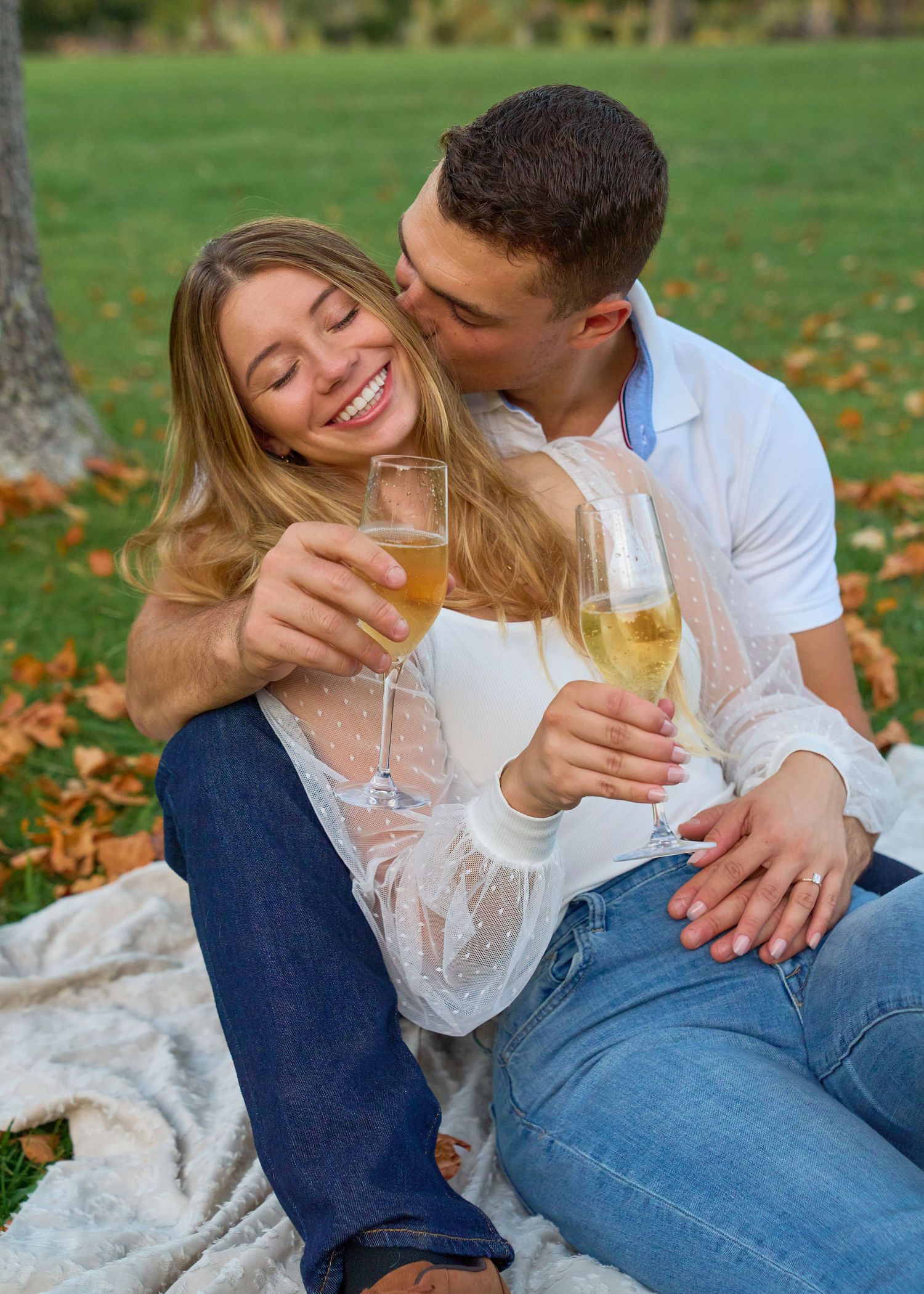 Smiling couple sharing champagne kiss on picnic blanket surrounded by autumn leaves in park.