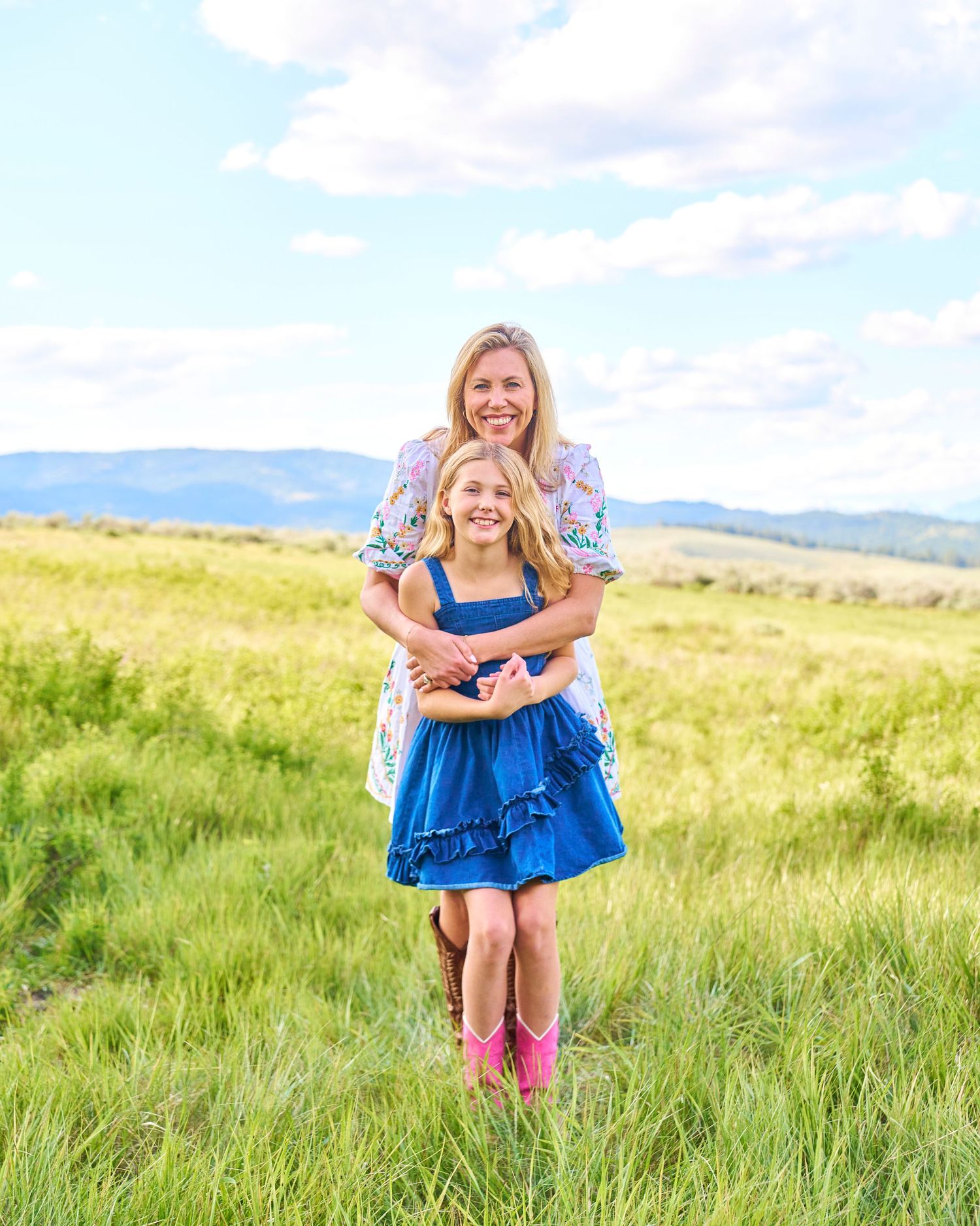 Smiling woman hugs girl in blue dress and pink boots standing in a bright green mountain meadow.