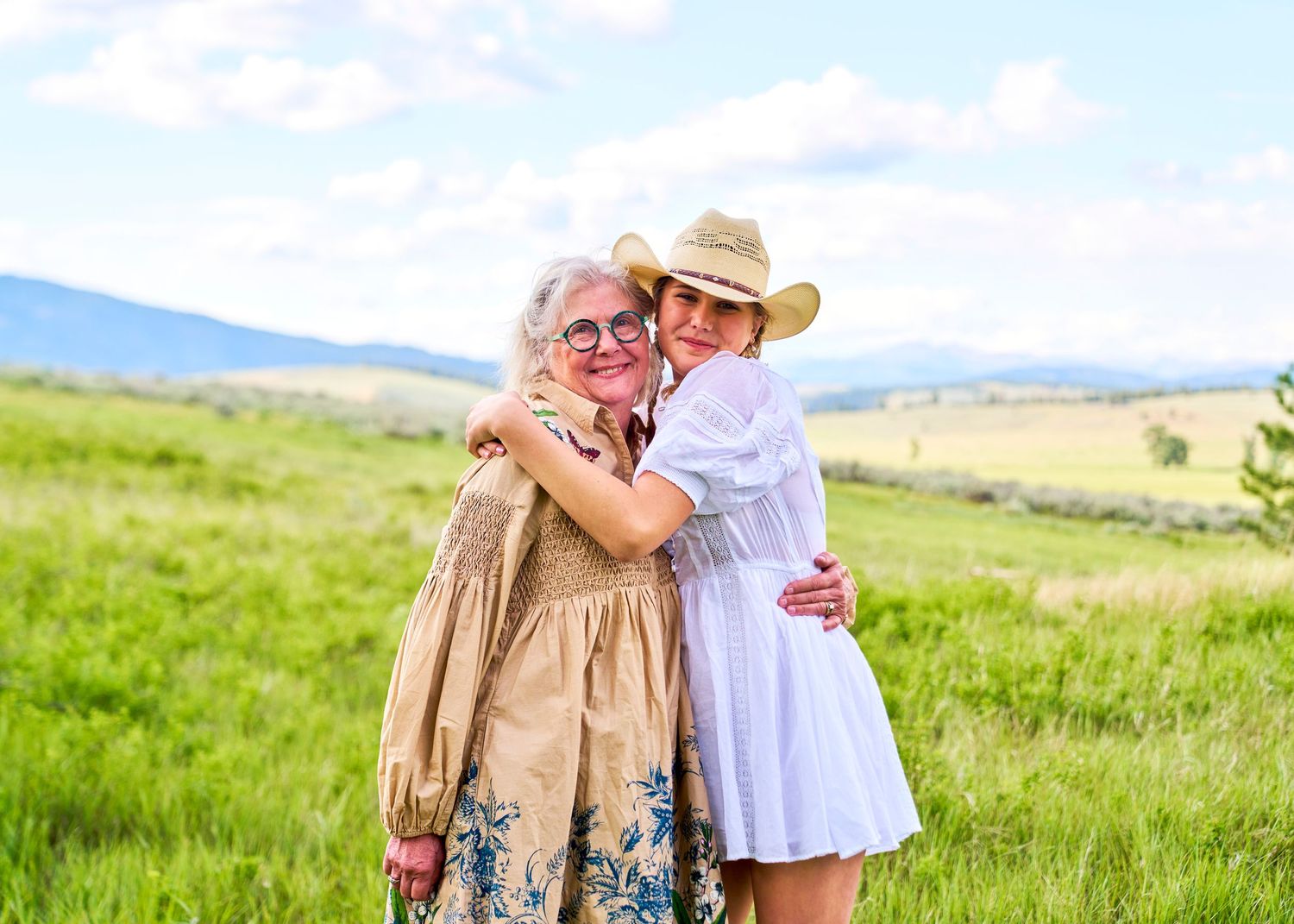 Two women hugging warmly in a sunny green meadow, both wearing straw hats and summer dresses.