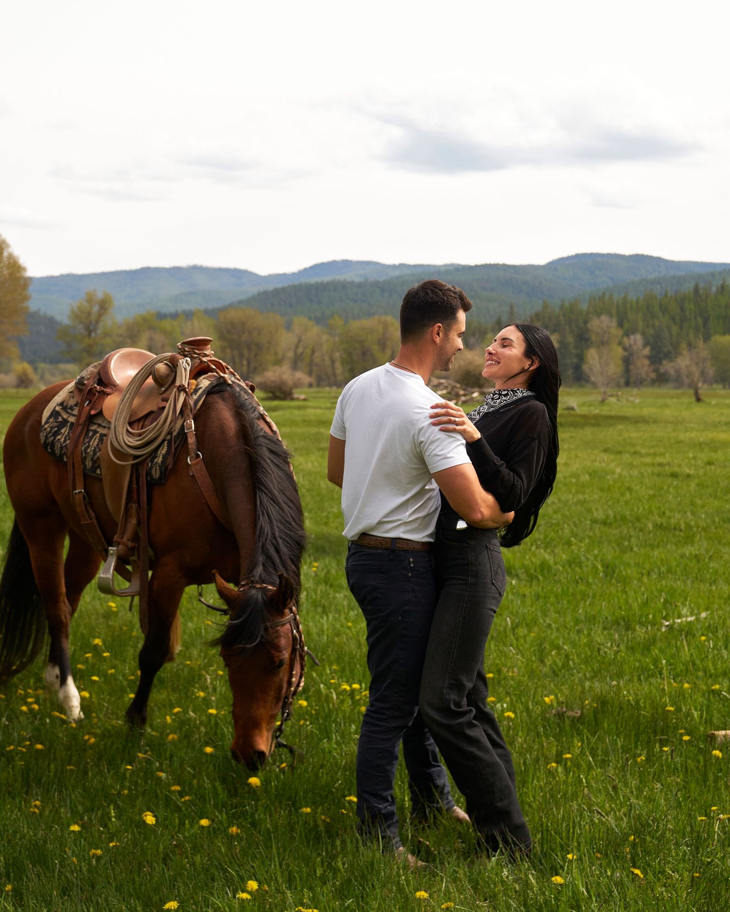 Couple sharing a tender moment in a green meadow beside a saddled brown horse with scenic mountain backdrop.