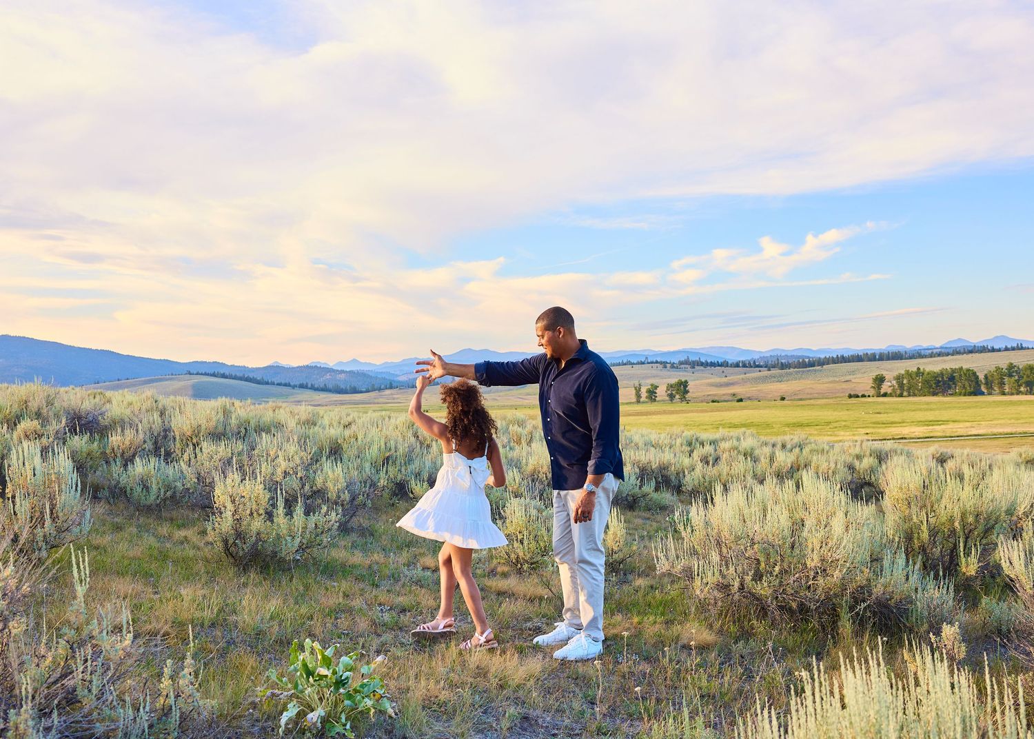 A father and daughter dance together in a wide open sagebrush meadow at sunset.