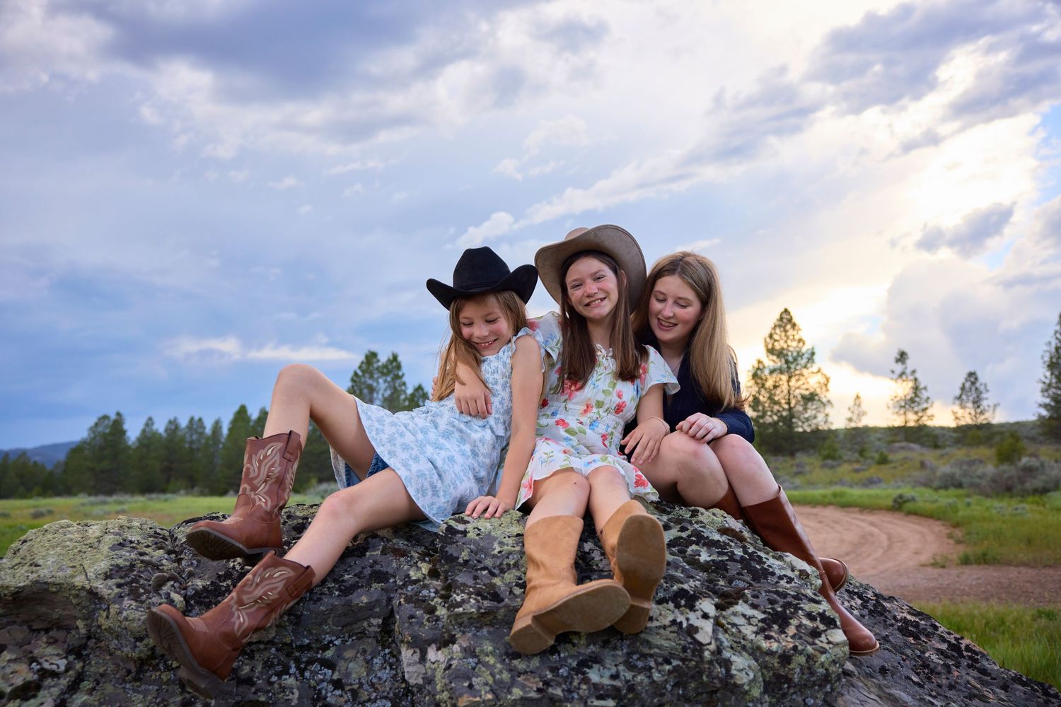 Three girls in floral dresses and cowboy boots sitting on a rock in a scenic outdoor setting.