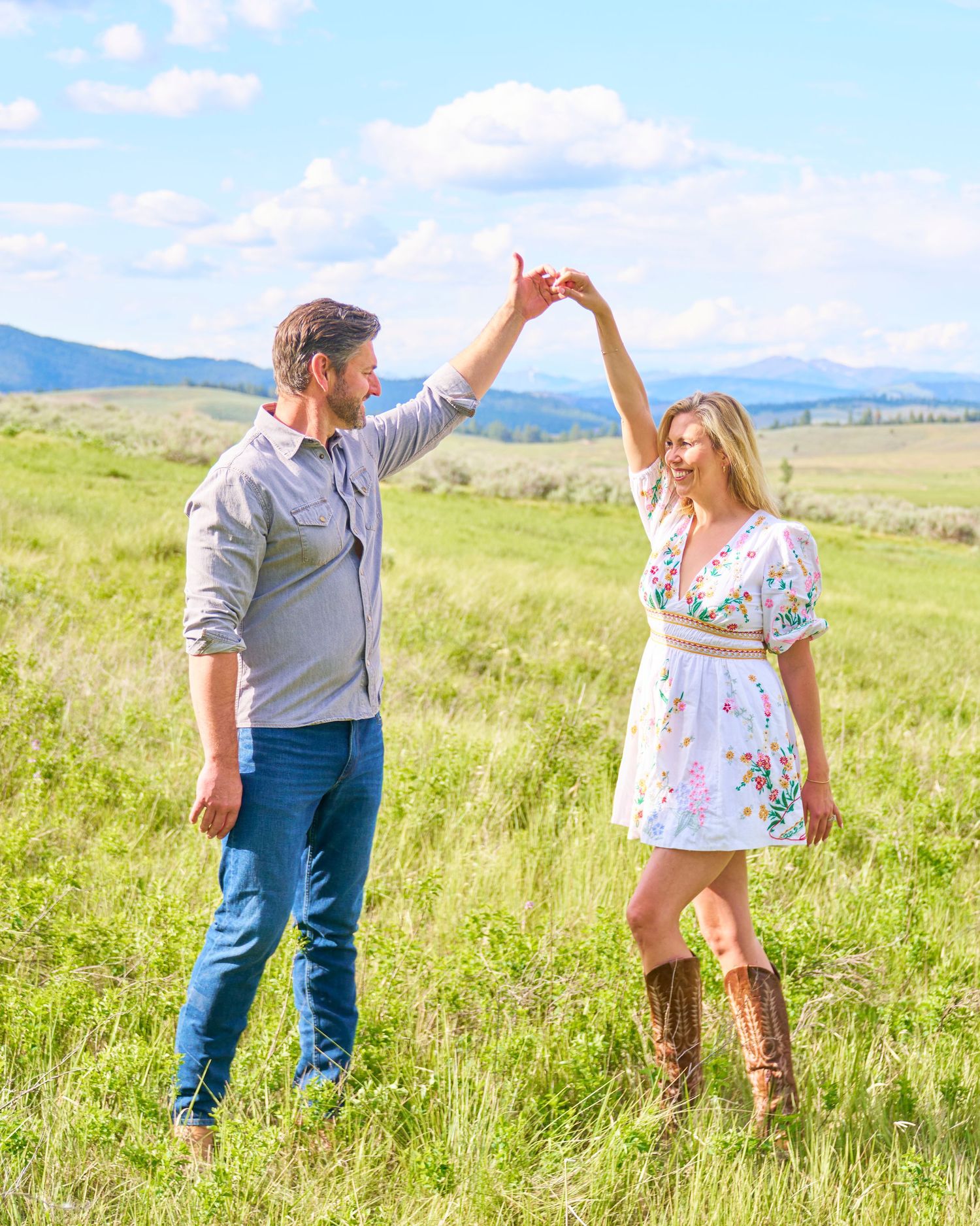 Couple dancing in sunny green meadow with mountains, man spinning woman in floral dress and boots.