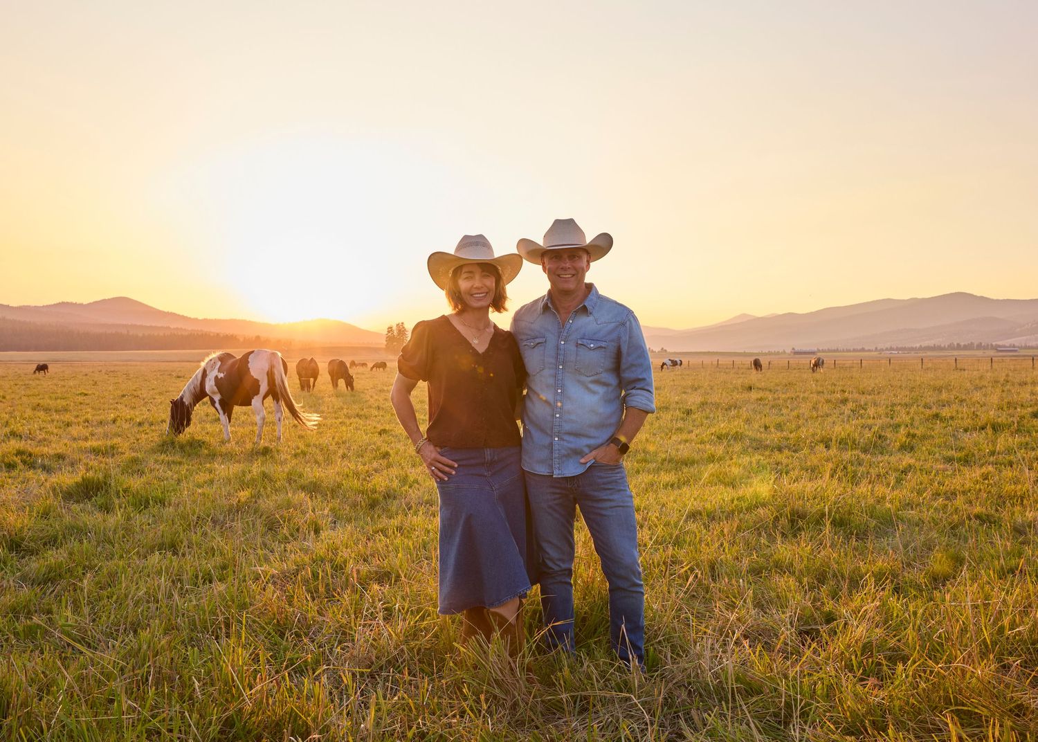 Couple in cowboy hats standing together in a ranch field at golden sunset.