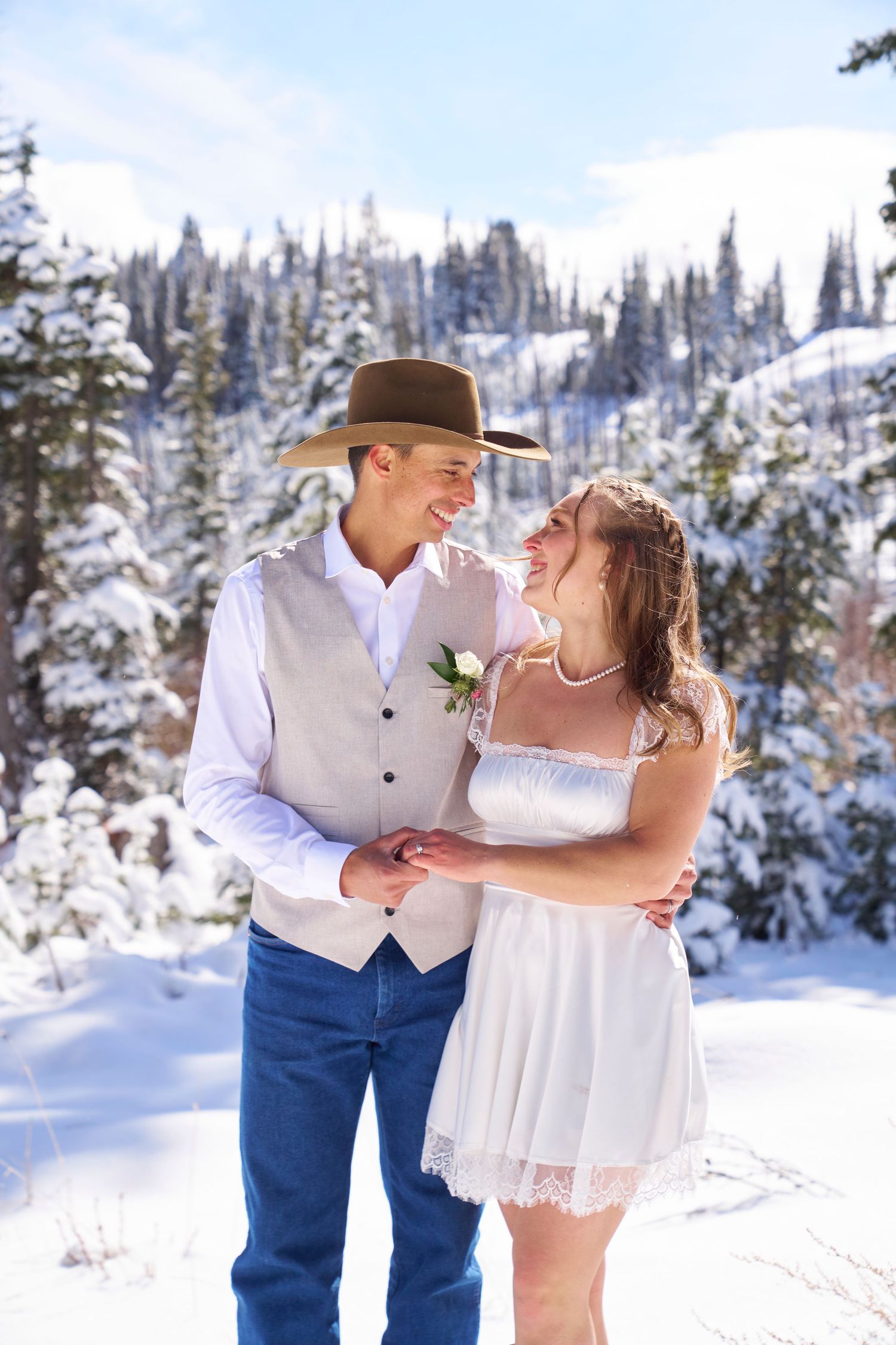 Couple in winter wedding attire embracing in snowy forest, groom in cowboy hat and vest, bride in white lace dress.
