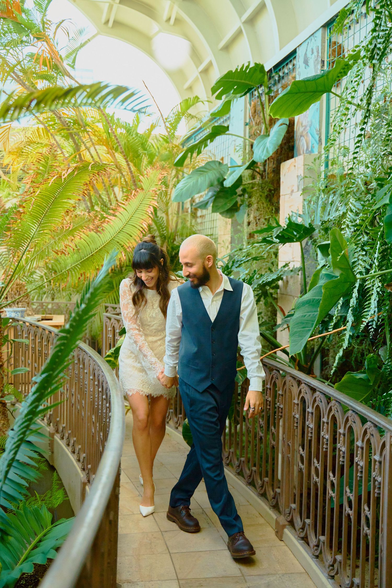 Newlyweds walk hand in hand through lush tropical greenhouse in wedding attire.