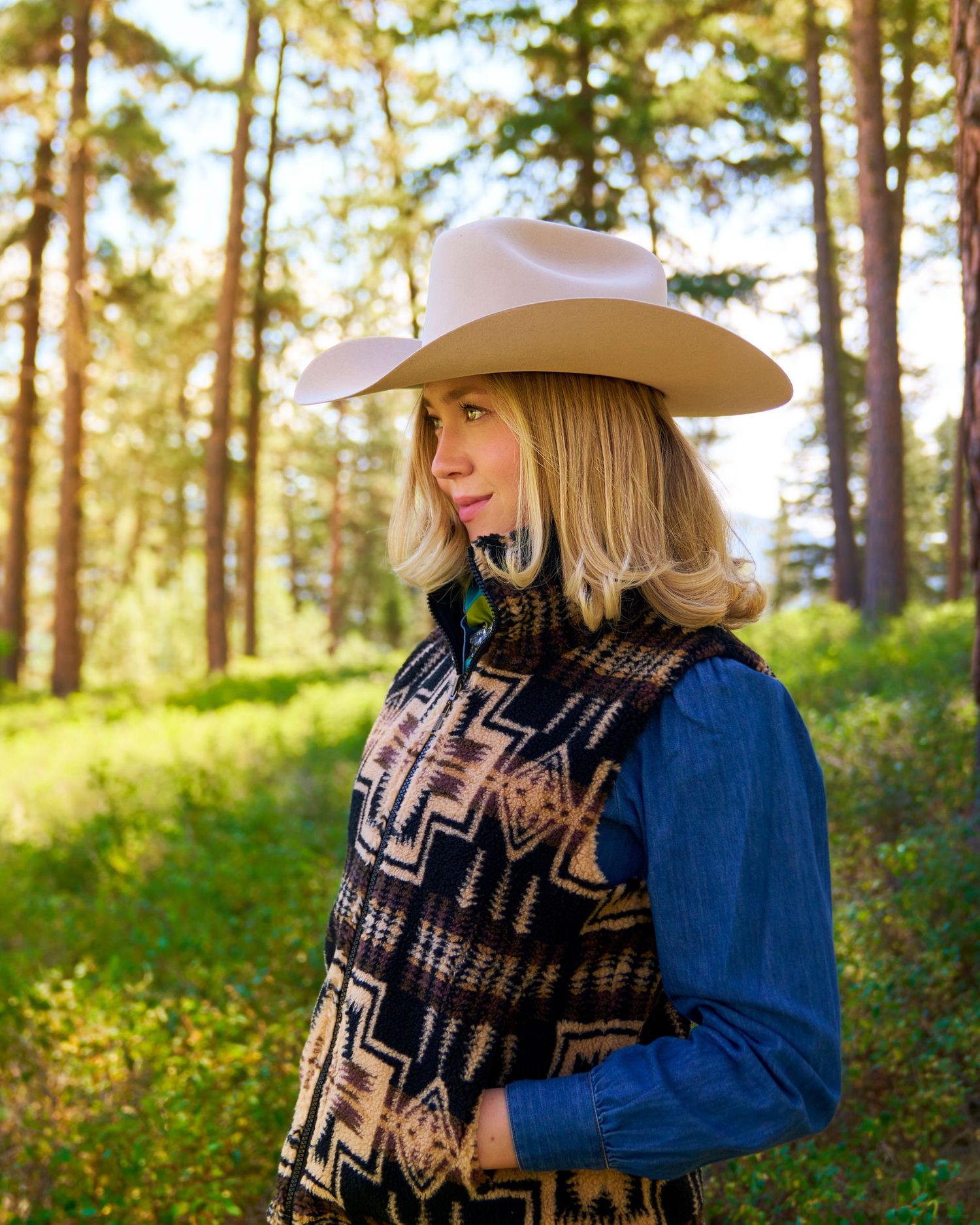 Blonde woman in a blue Pendleton dress and Stetson hat poses among tall pine trees in a sunlit forest.