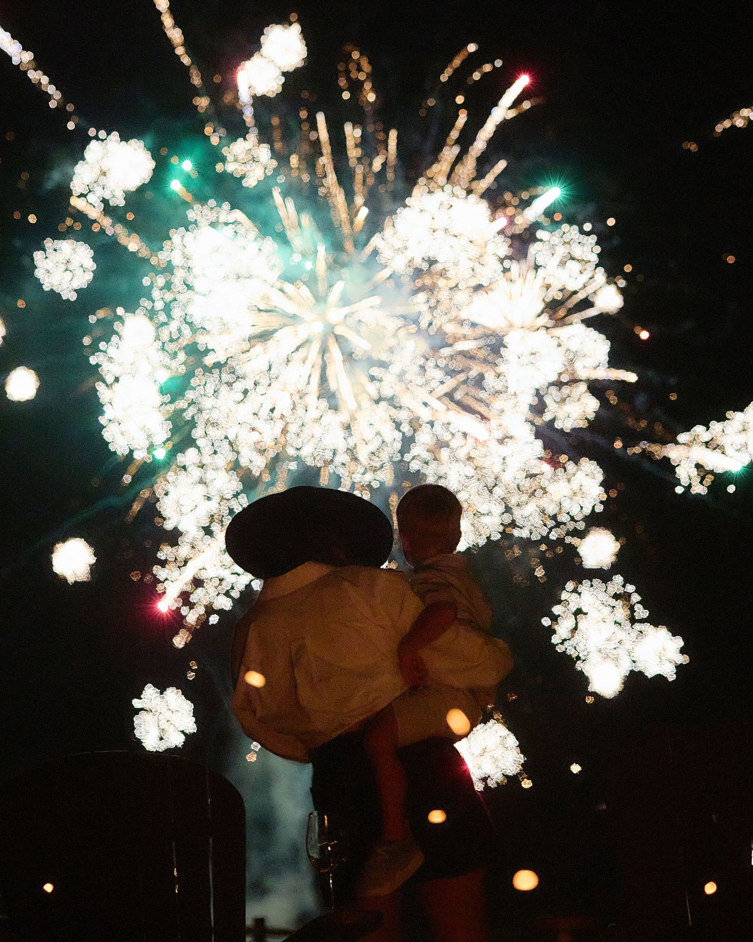 Silhouette of person holding a child in a sombrero watching brilliant white fireworks burst in the night sky.