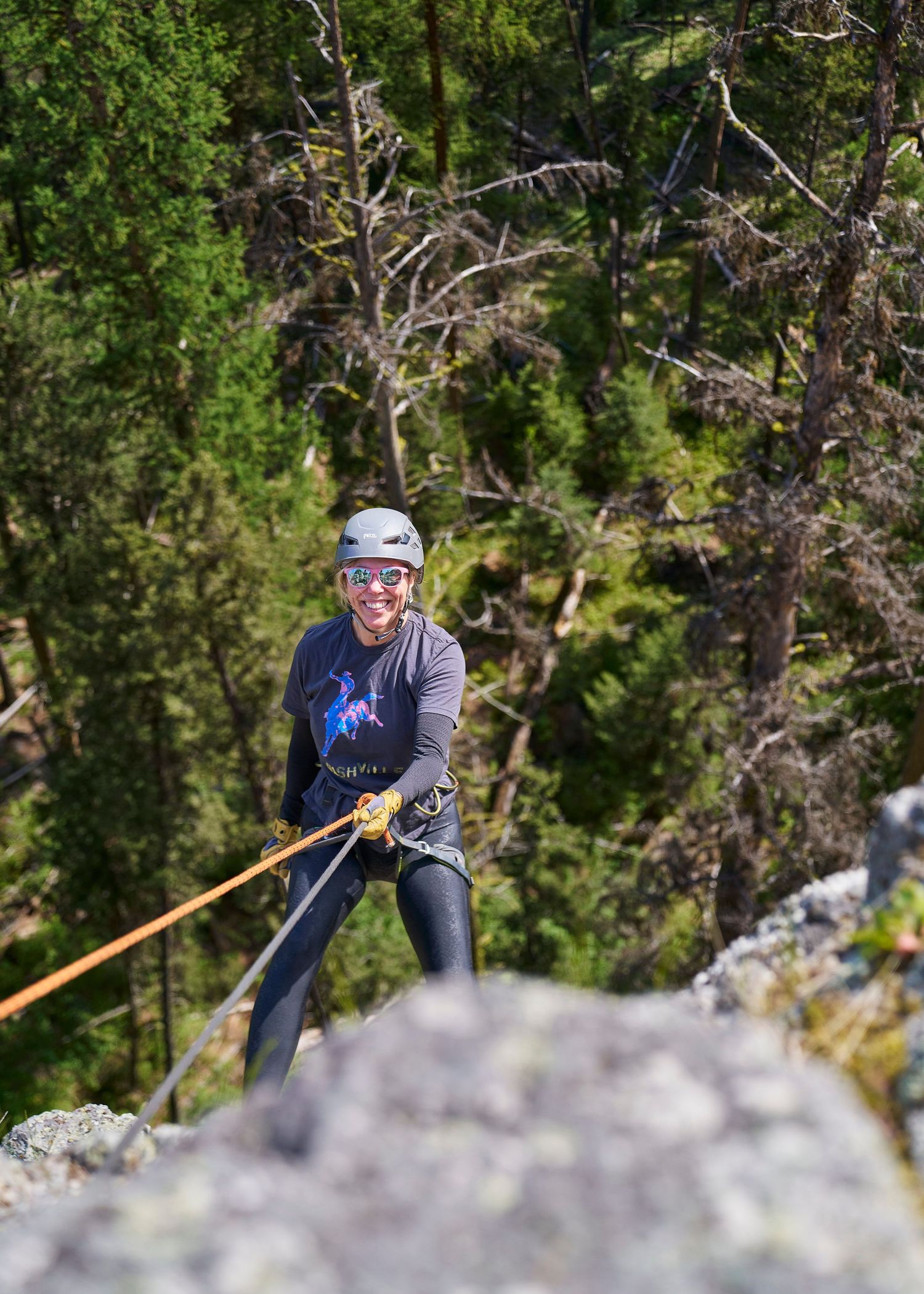 A smiling woman rappelling down a rocky cliff face surrounded by green forest trees.