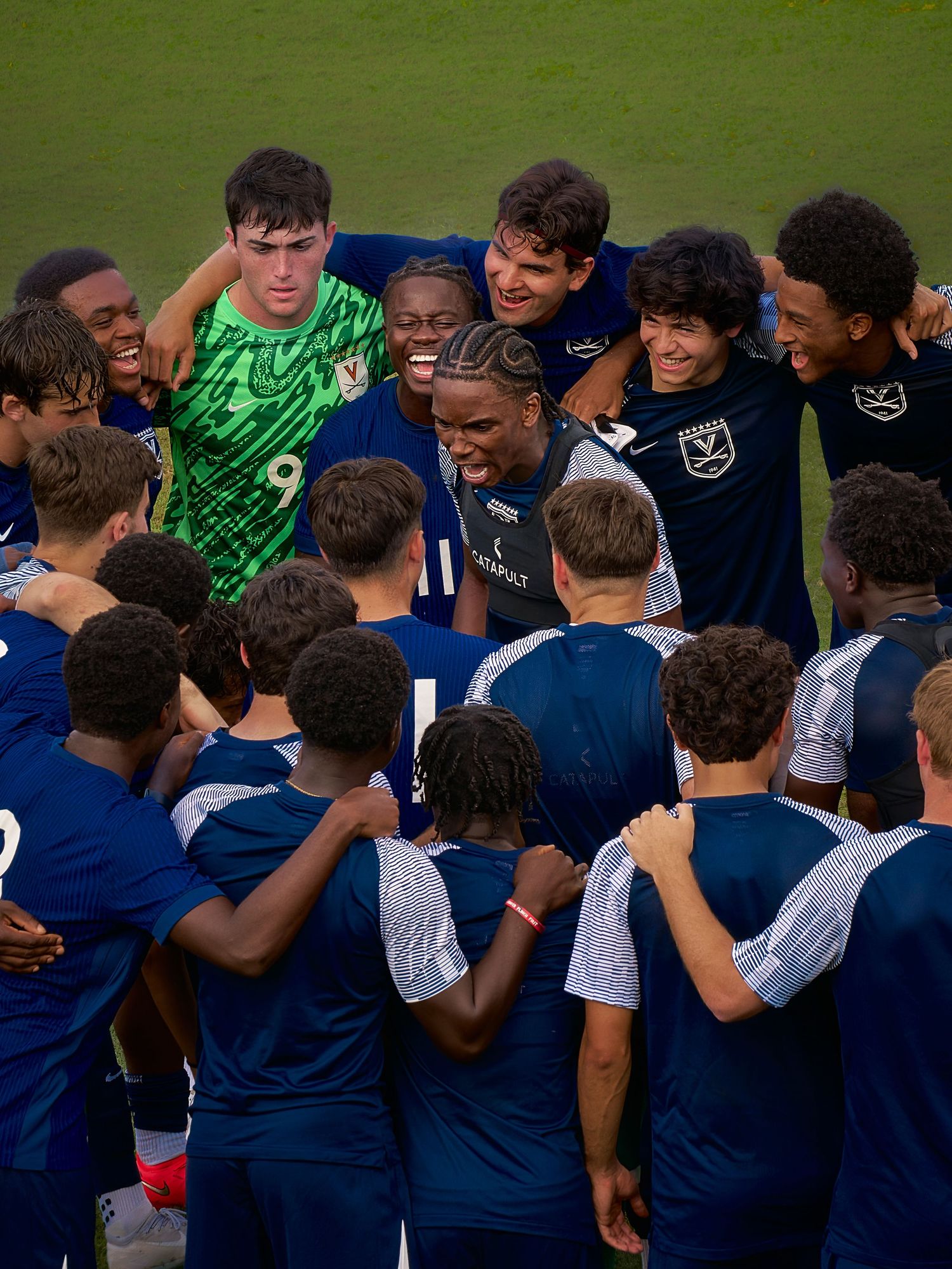 Youth soccer team huddles together in navy blue uniforms, celebrating with smiles on a green field.