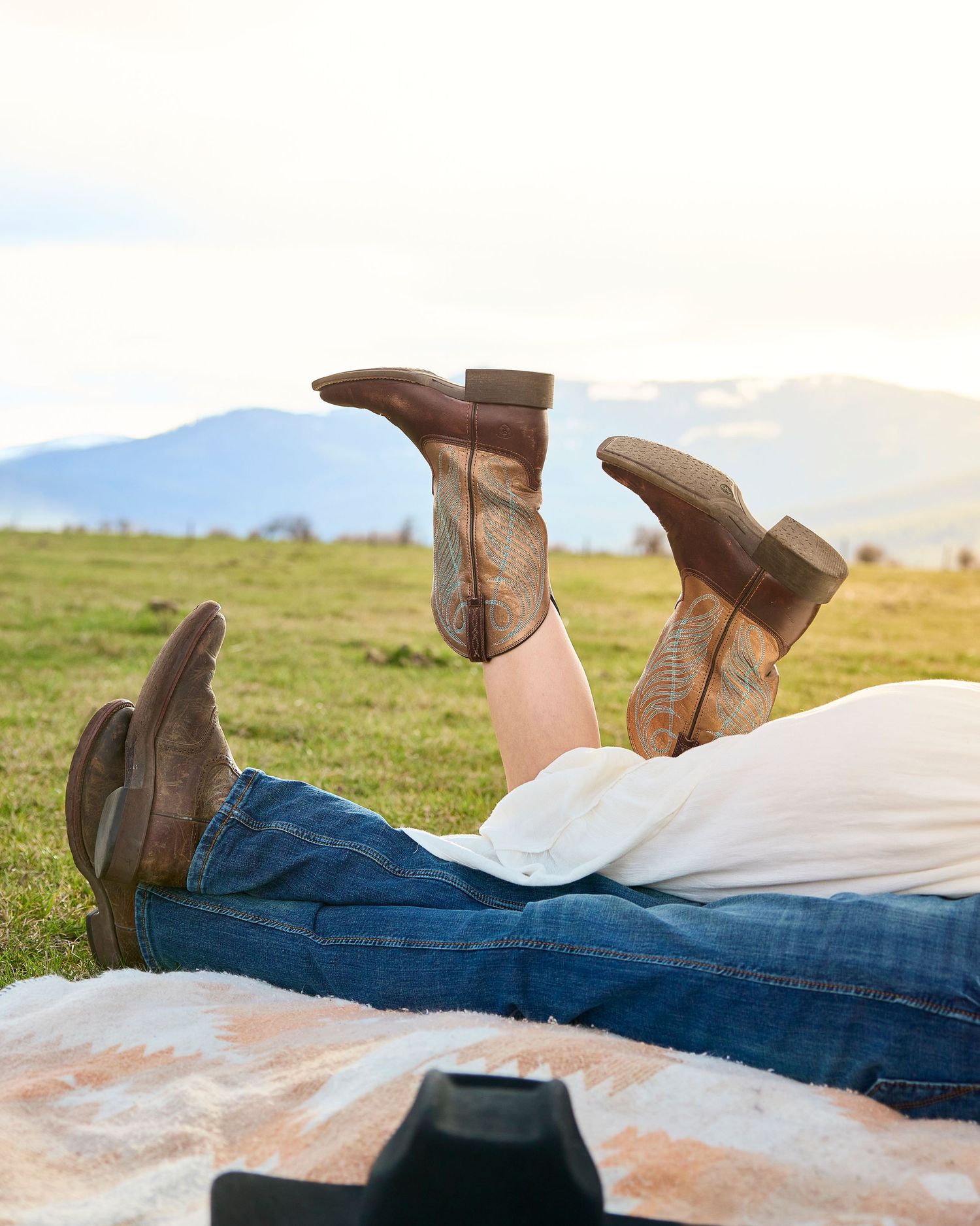 Couple lies in grass with cowboy boots raised, mountain landscape visible in background.
