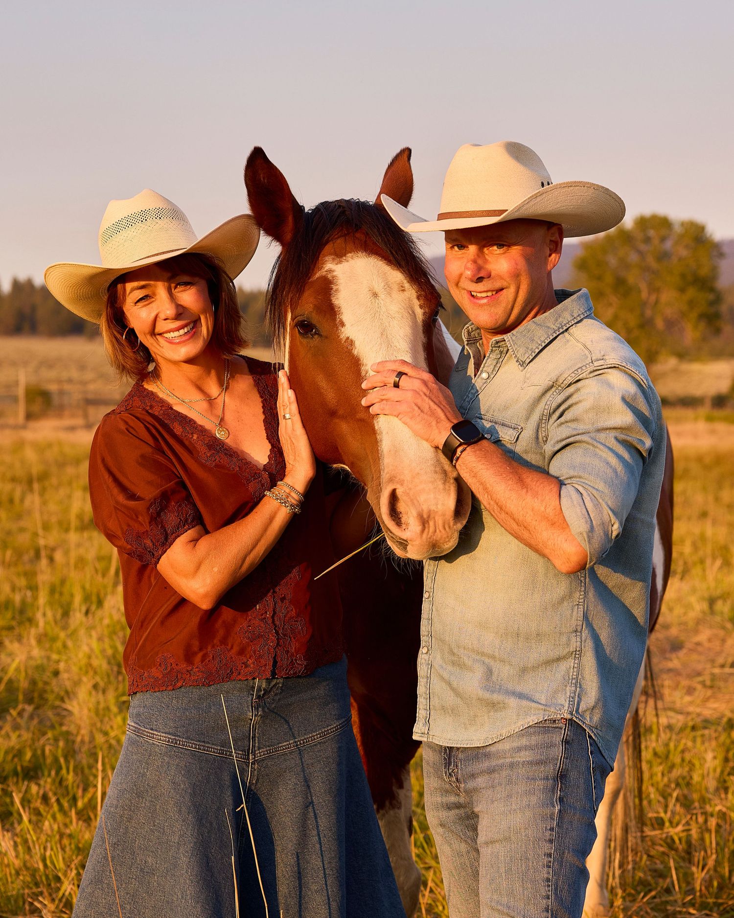 Smiling couple in white cowboy hats posing with paint horse in sunny ranch pasture.