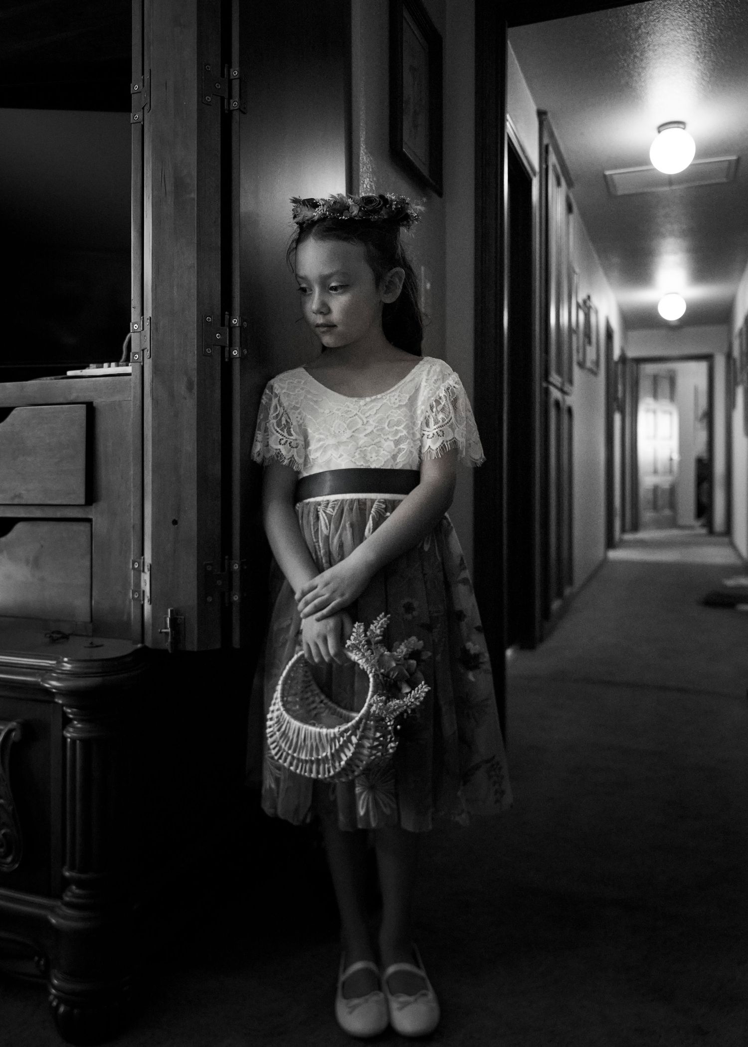 Young girl in lace dress holding decorative purse stands in dimly lit hallway in black and white photo.