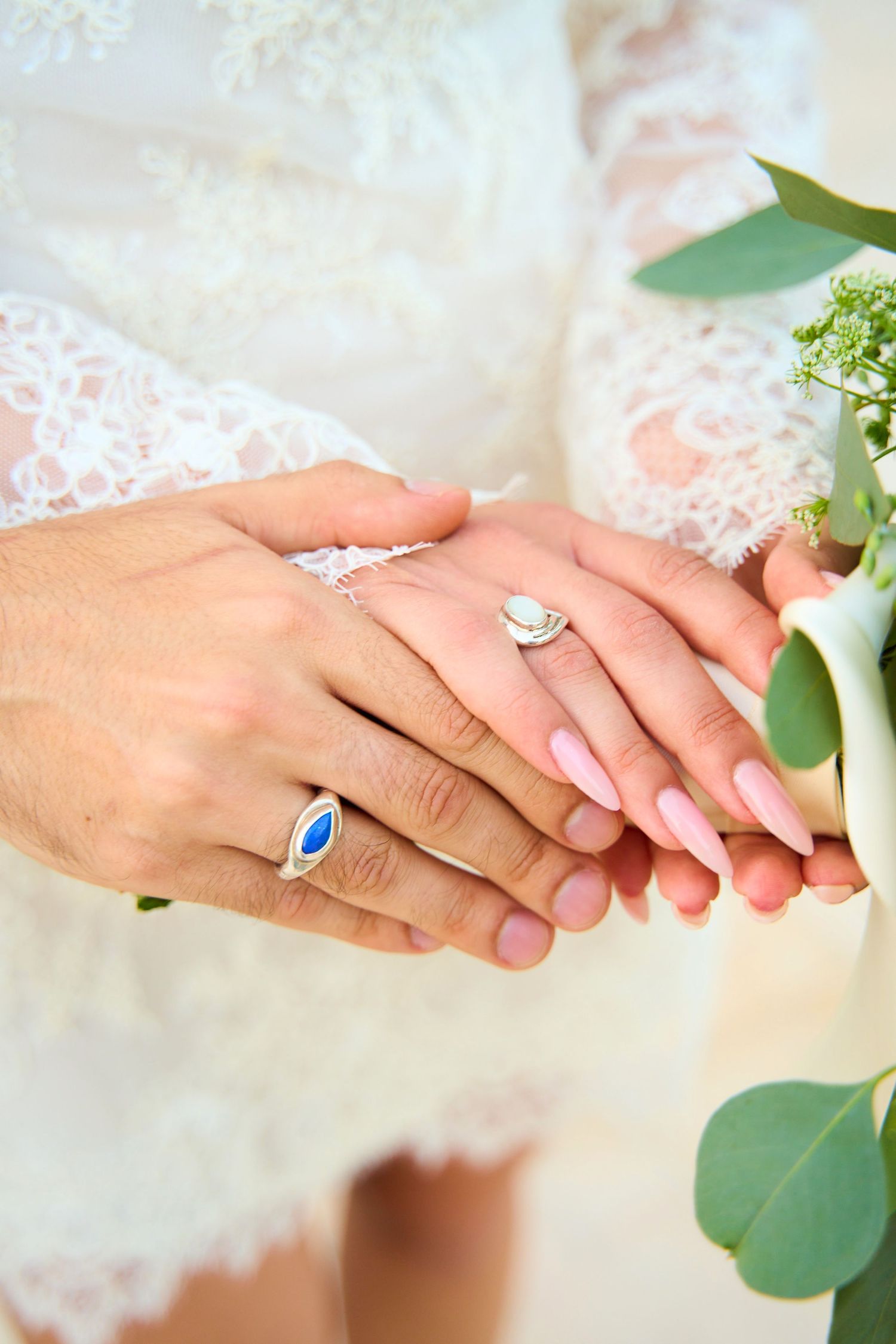 Close-up of bride and groom hands showing wedding rings, with white lace dress and bridal bouquet.