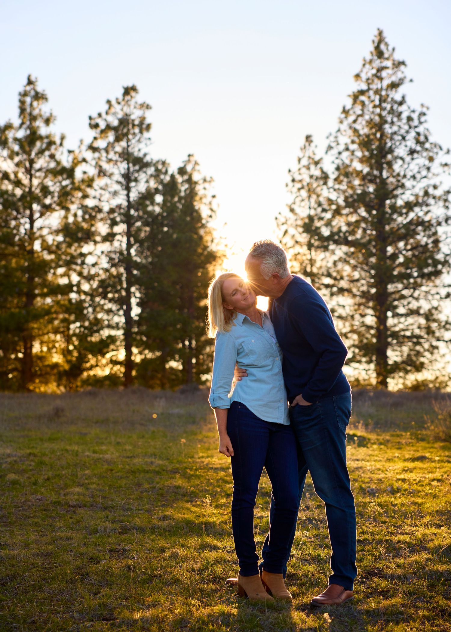 Couple sharing a romantic golden hour kiss in a sunlit field with tall pine trees in the background.
