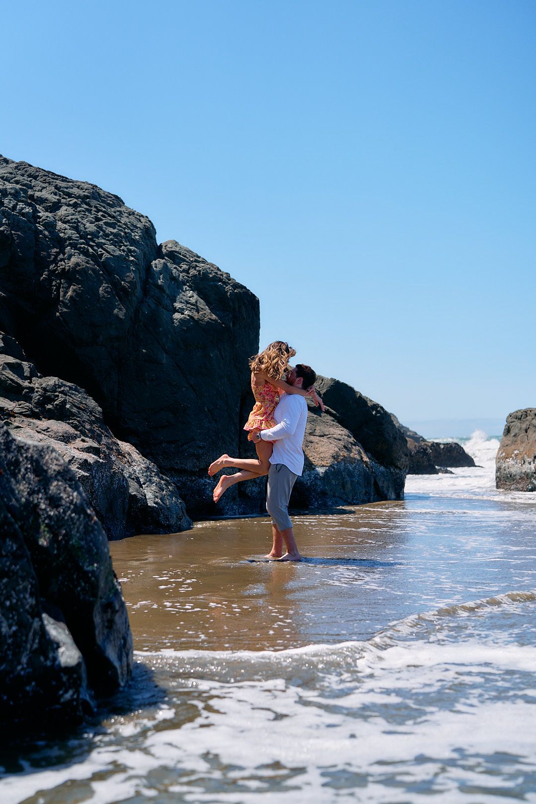 A couple shares a romantic kiss on a rocky beach, man lifting woman near ocean waves on a sunny day.