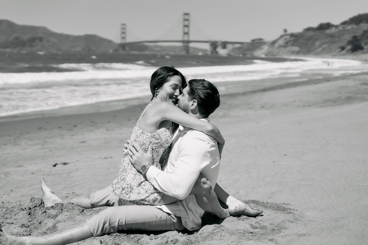 Black and white photo of couple sitting intimately on Baker Beach near Golden Gate Bridge.
