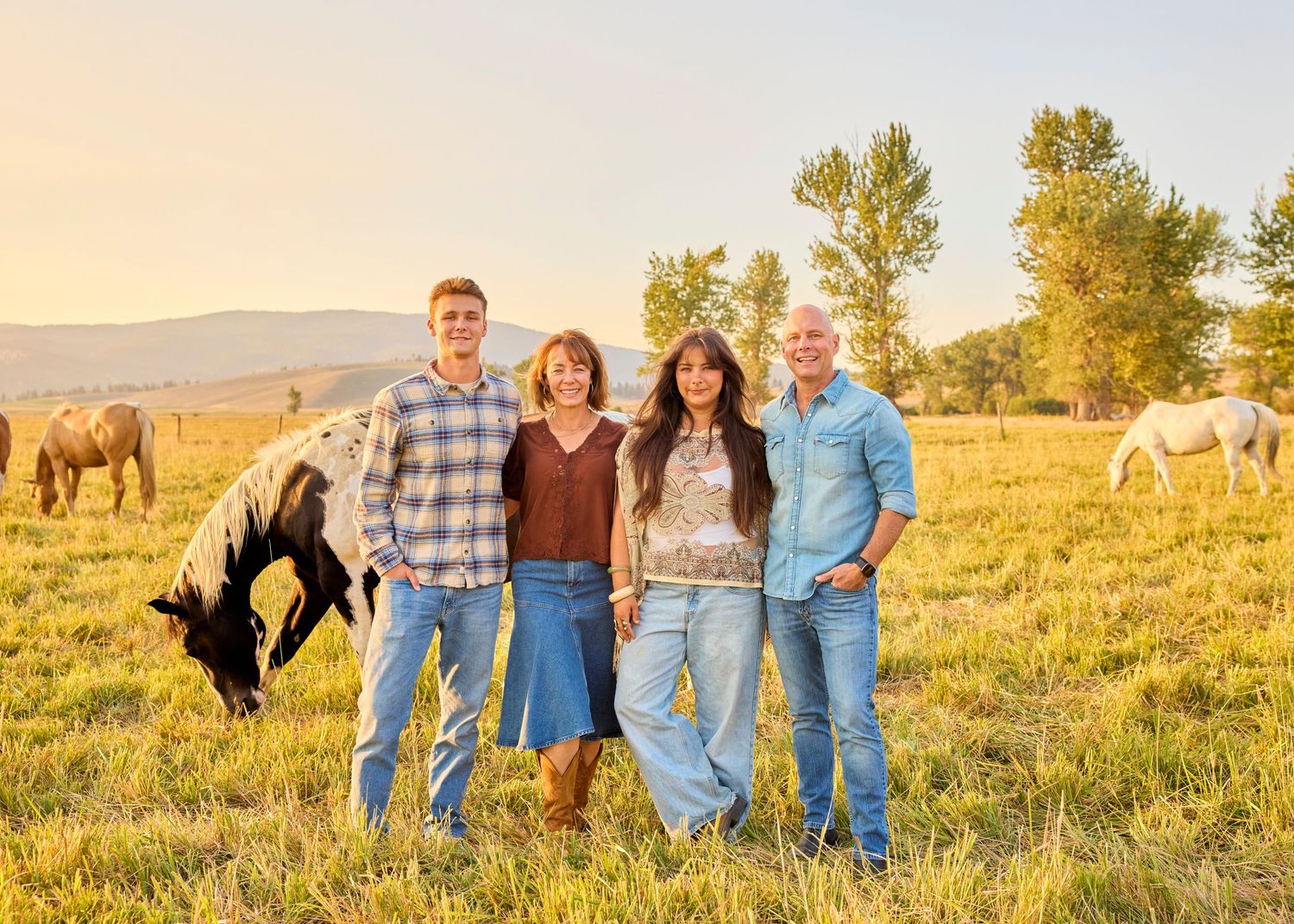 Family of four smiling together in green pasture with horses grazing at golden hour.