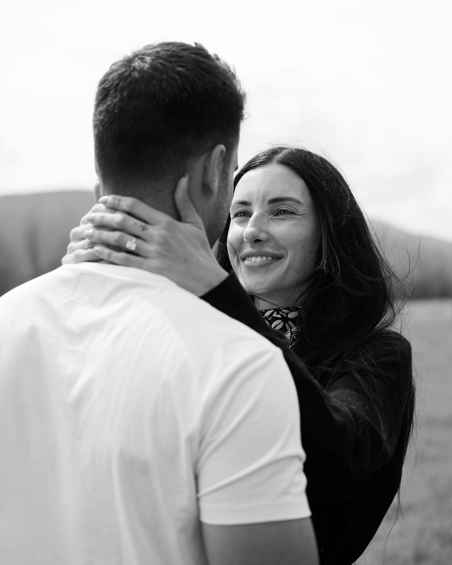 Black and white close-up of smiling dark-haired woman embracing man outdoors in an open field.