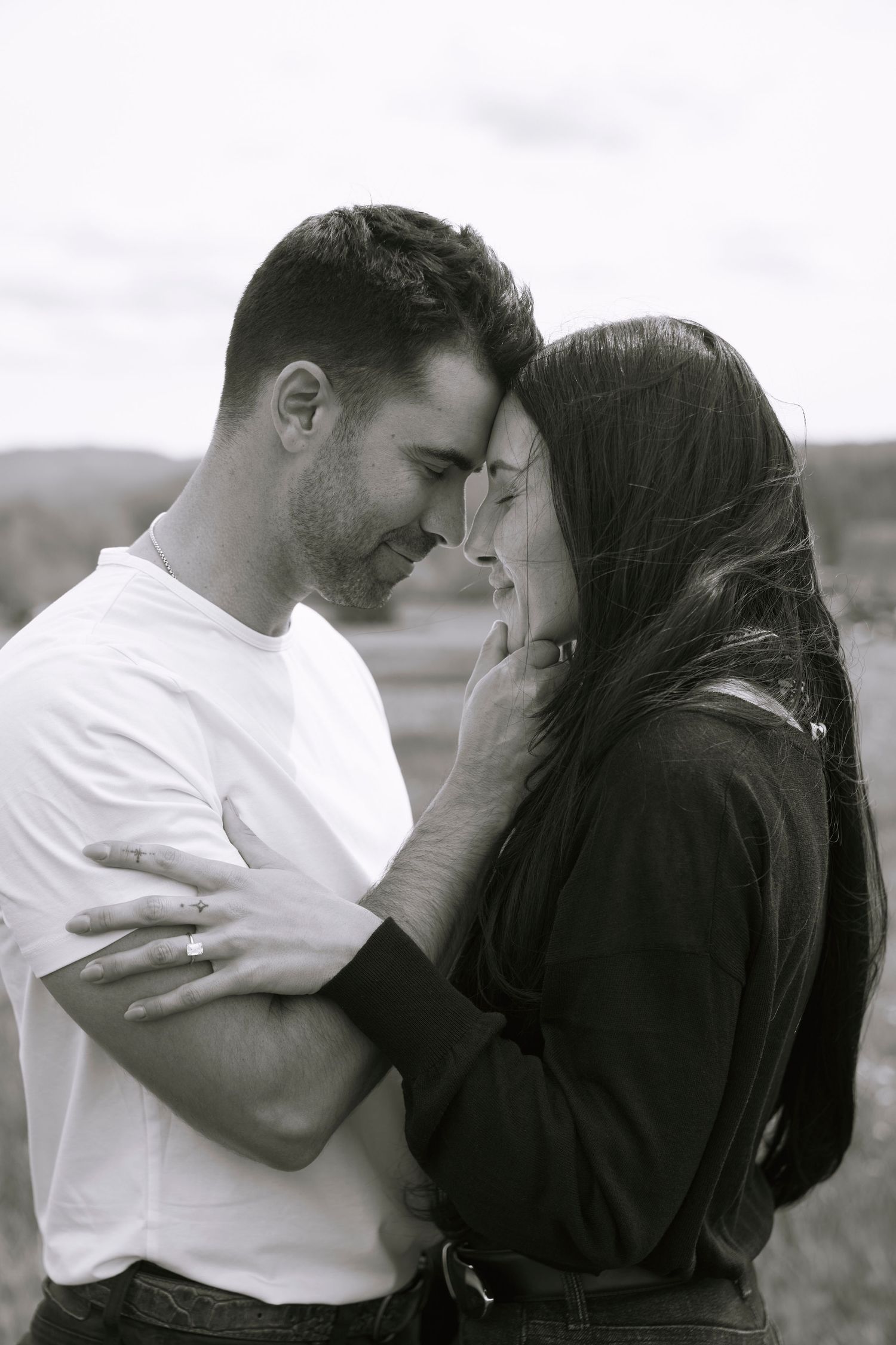Black and white photo of a romantic couple embracing closely outdoors, touching foreheads tenderly.