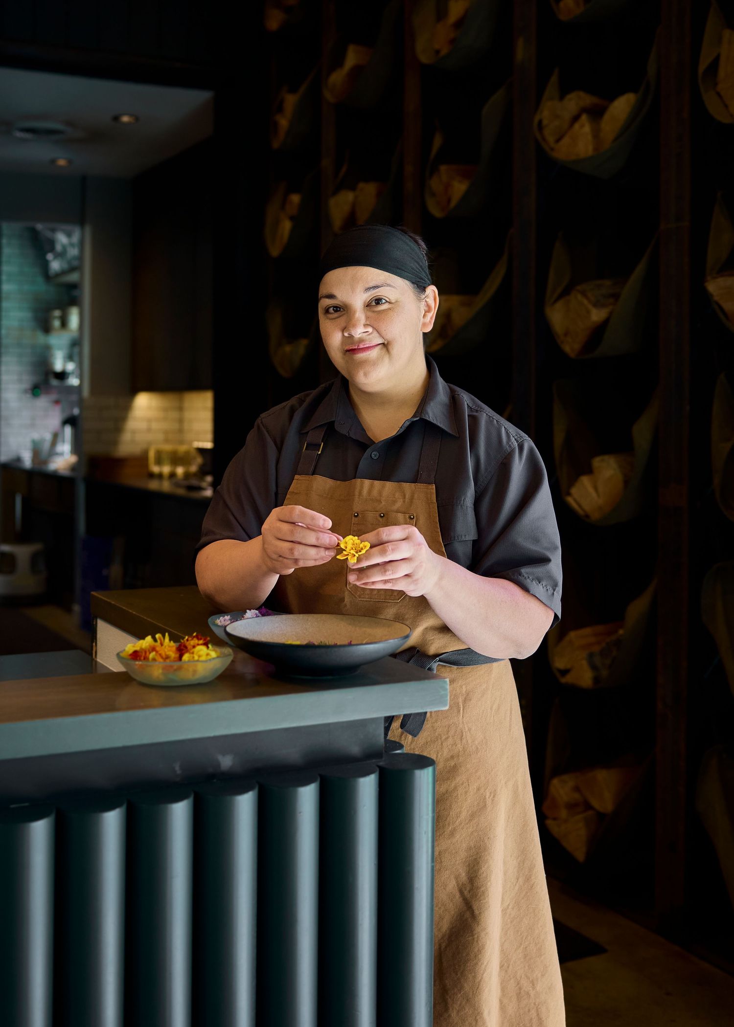 Female chef in black cap and apron plating colorful dish at restaurant counter.