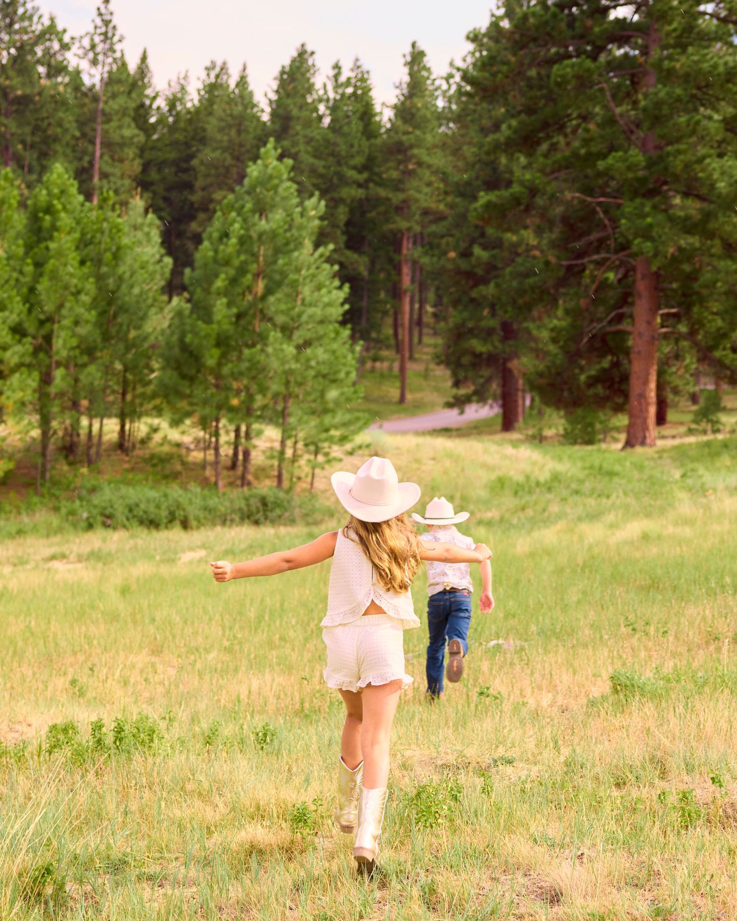 Two children in cowboy hats running through a lush green meadow surrounded by pine trees.