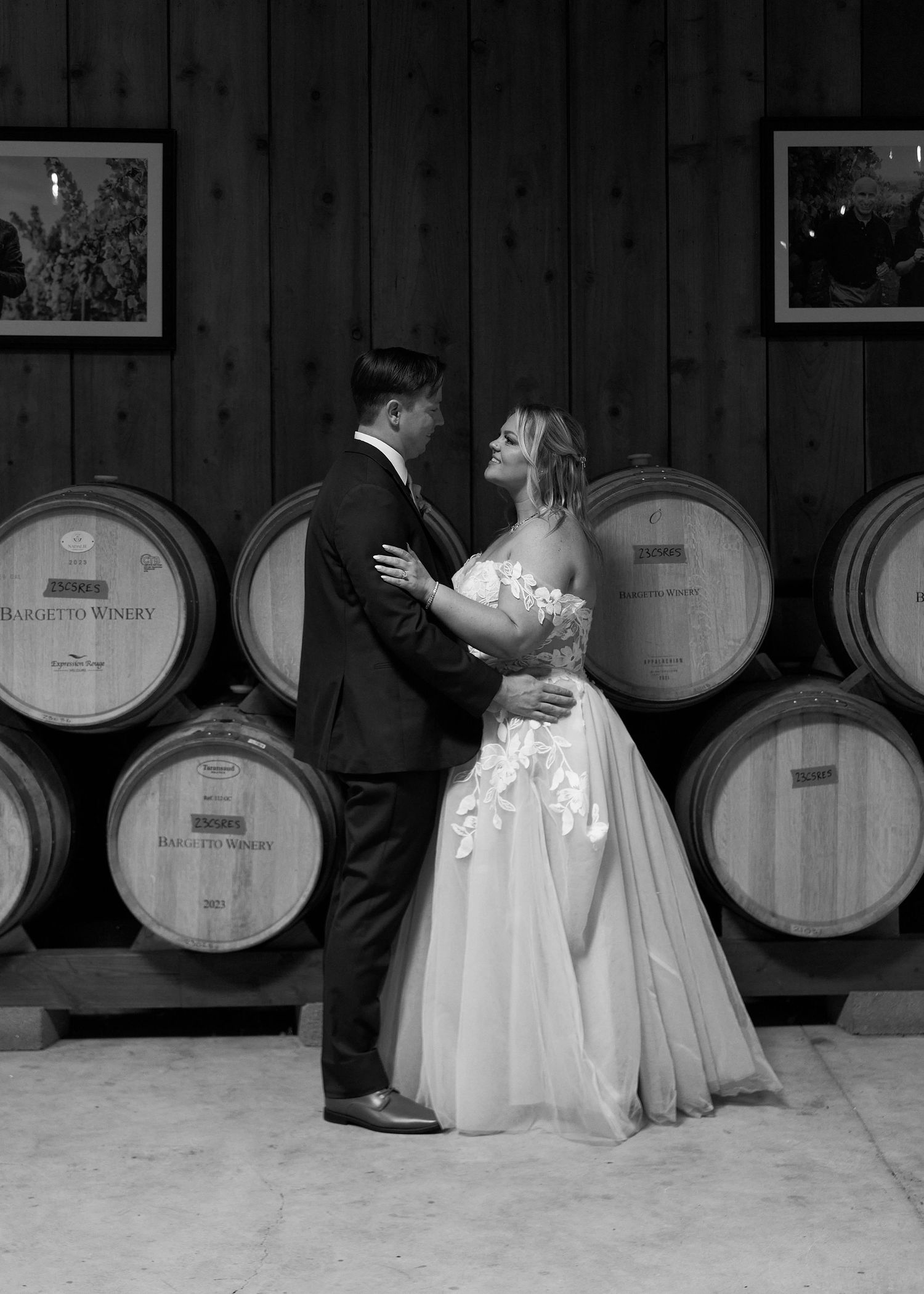 Black and white photo of bride and groom embracing before rustic winery barrel wall.