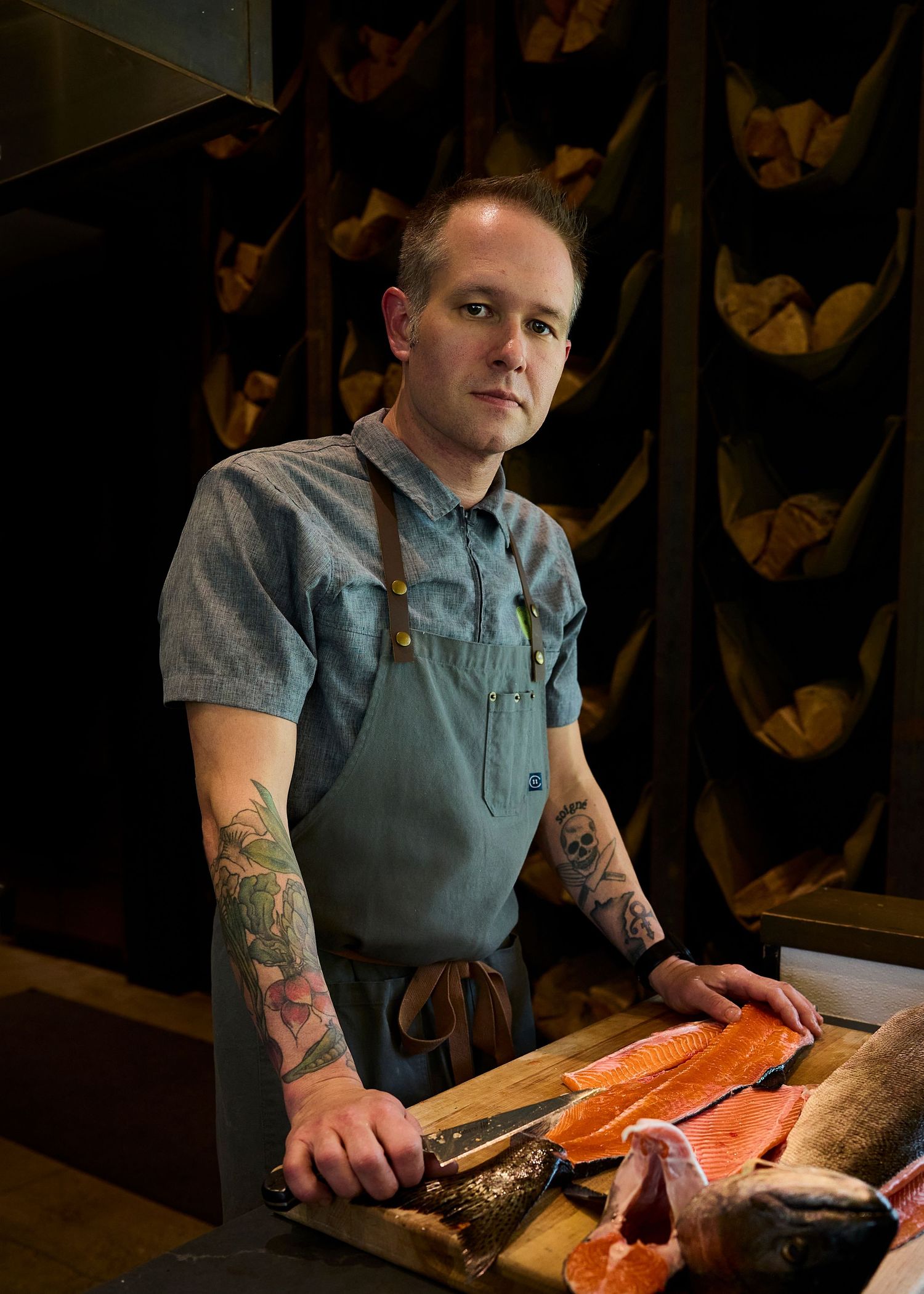 Tattooed chef in blue shirt prepares fresh salmon fillet on wooden cutting board in dimly lit restaurant kitchen.