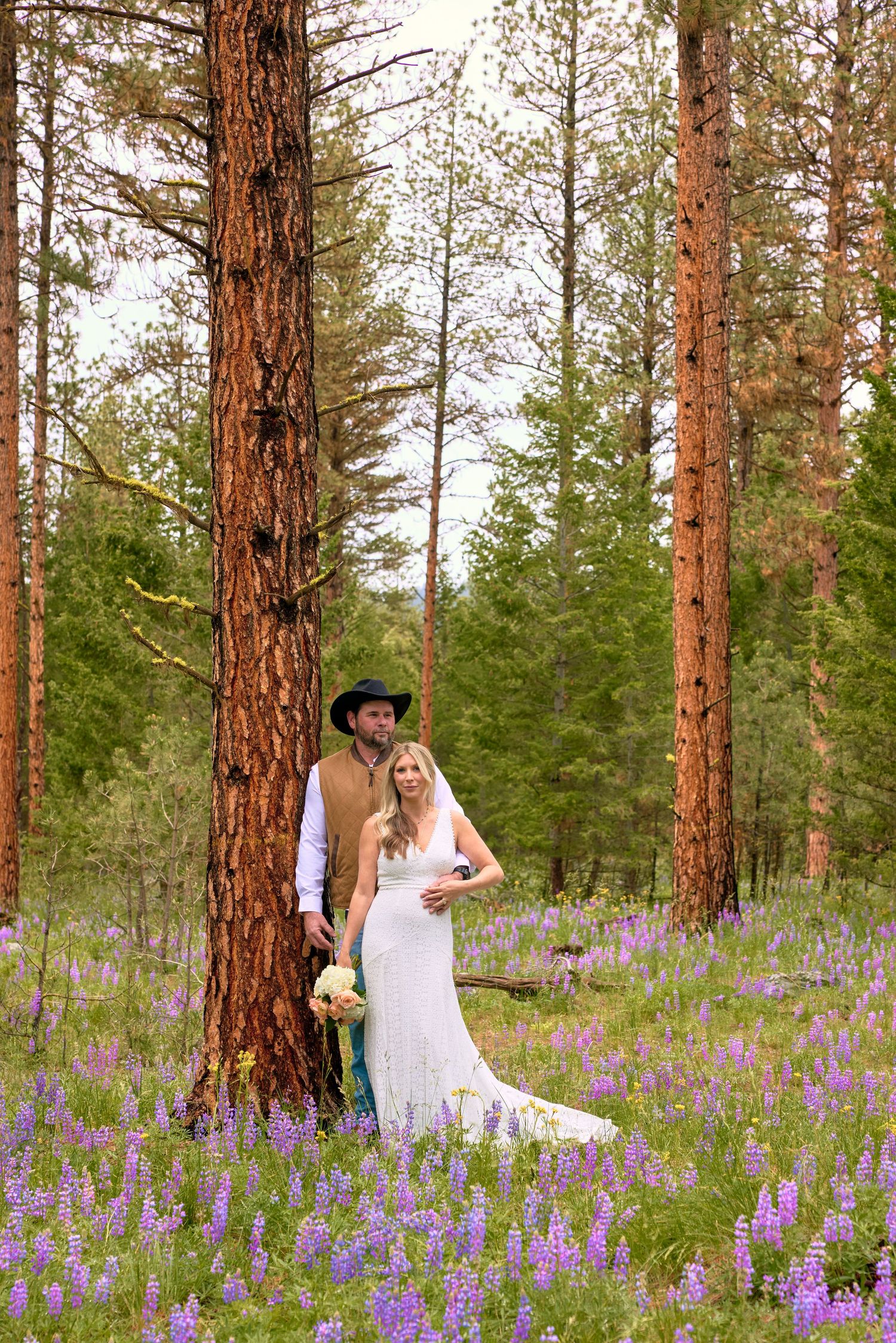 Bride and groom pose by a tall pine tree in a forest meadow filled with purple lupine flowers.