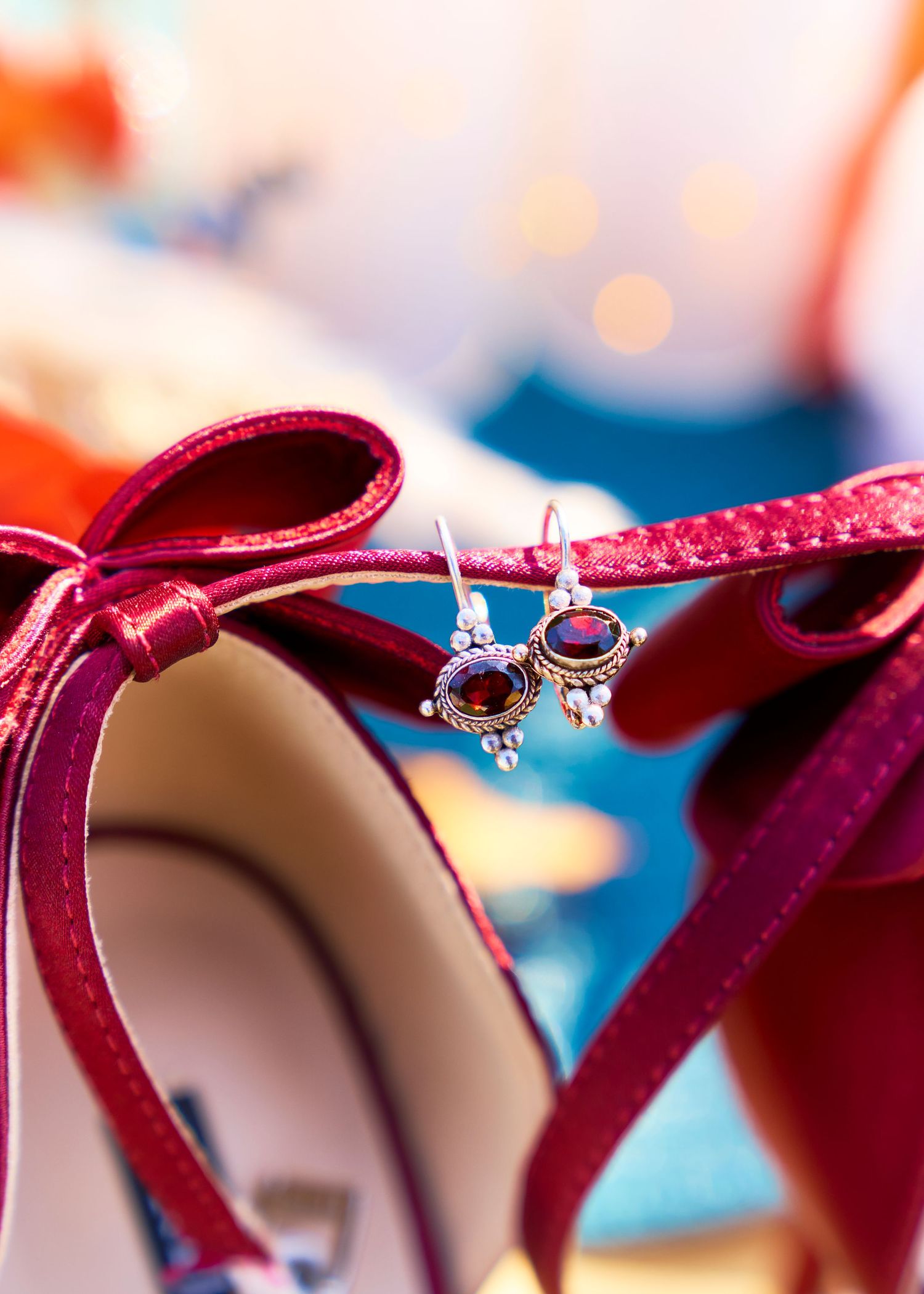 Red satin bridal heels with bow detail and dangling garnet earrings displayed as elegant wedding accessories.