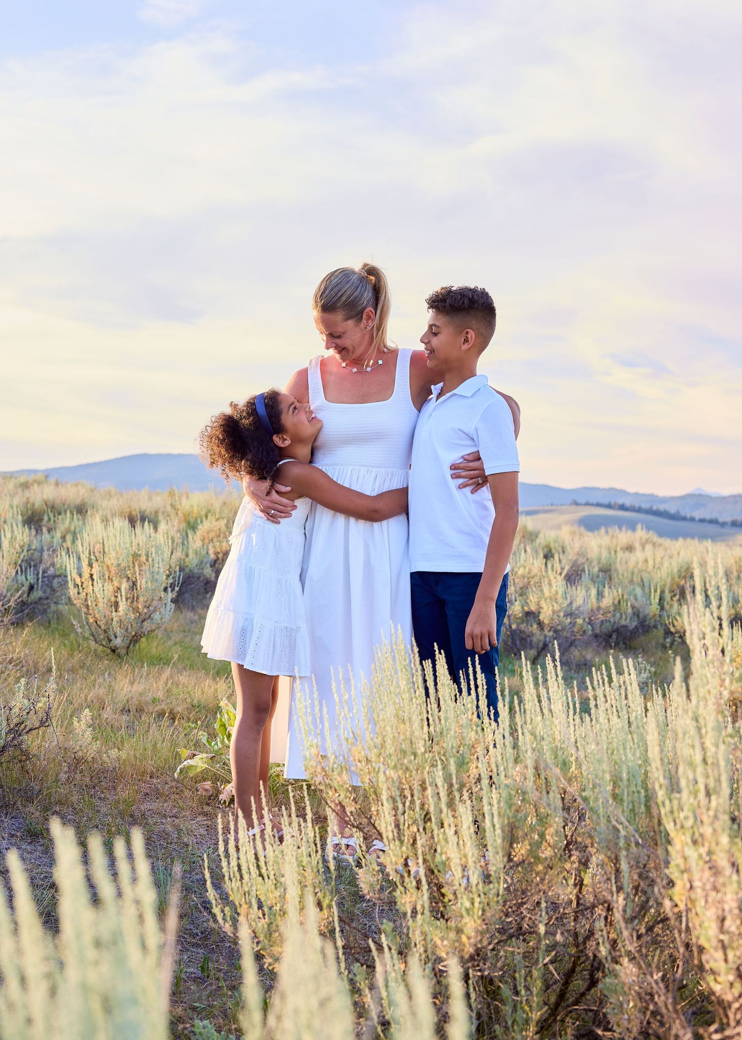 Mother in white dress laughing with two children in sagebrush meadow at sunset.
