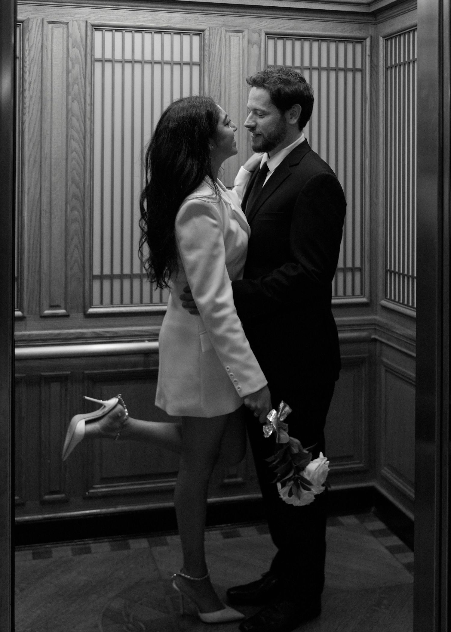 Black and white photo of a couple sharing a romantic moment in an elevator, woman in white blazer dress lifting one heel.