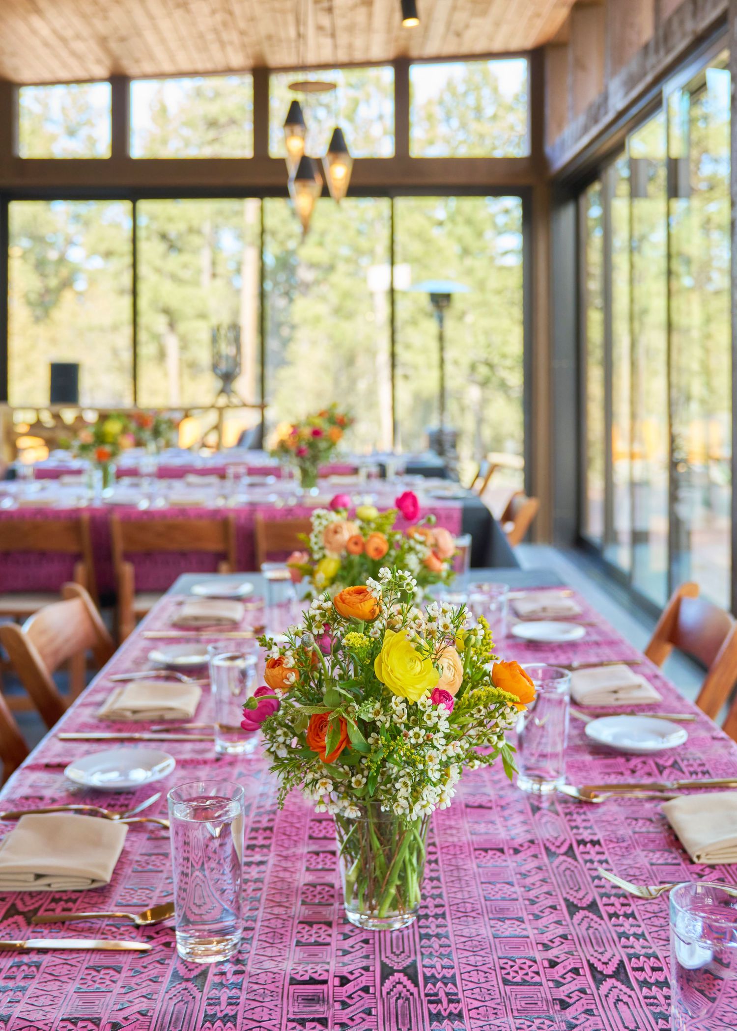 Elegant indoor dining table set with pink patterned linens and fresh wildflower centerpieces in a glass venue.