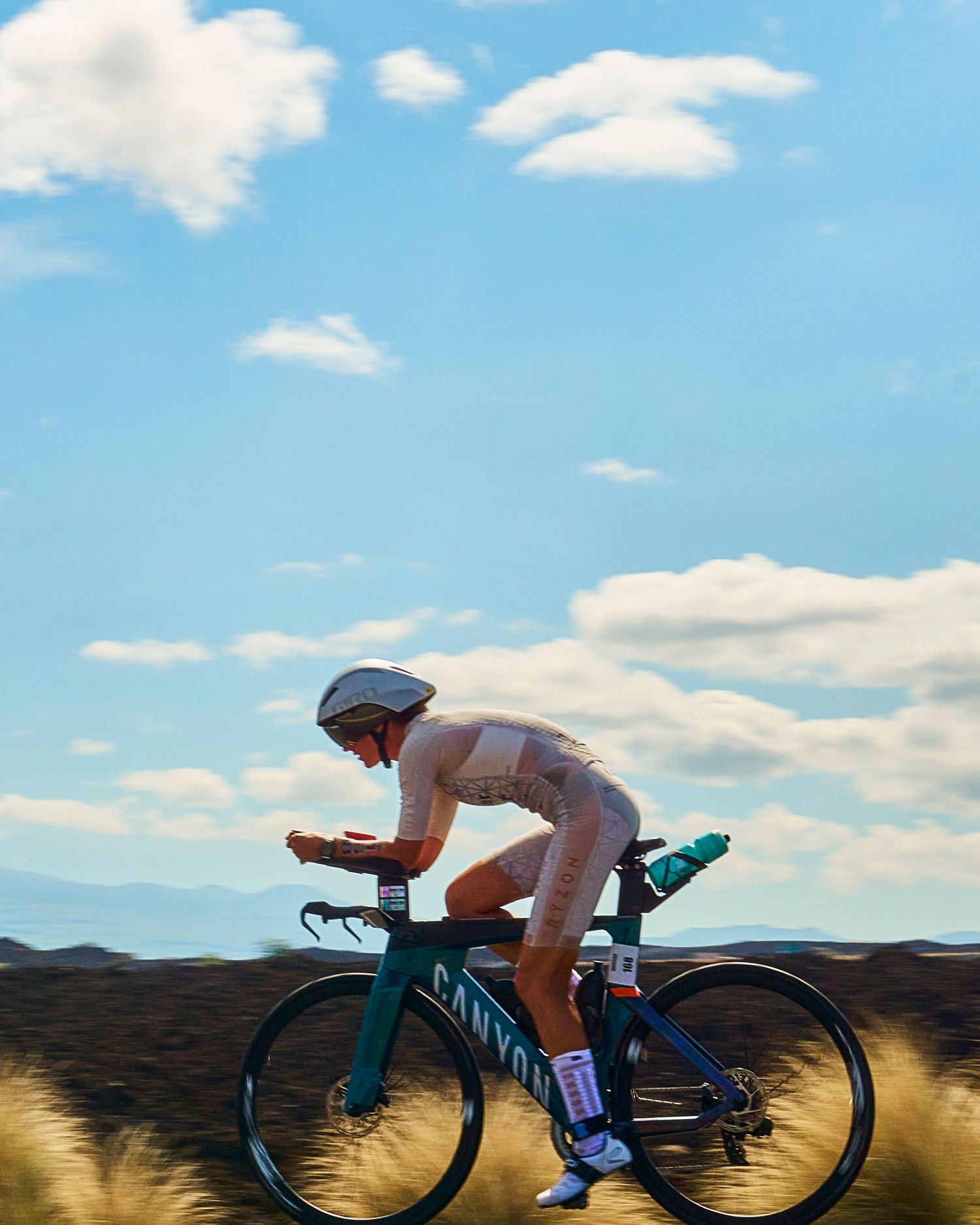 Cyclist in aerodynamic position riding a Canyon triathlon bike under a blue sky with clouds in a desert landscape.