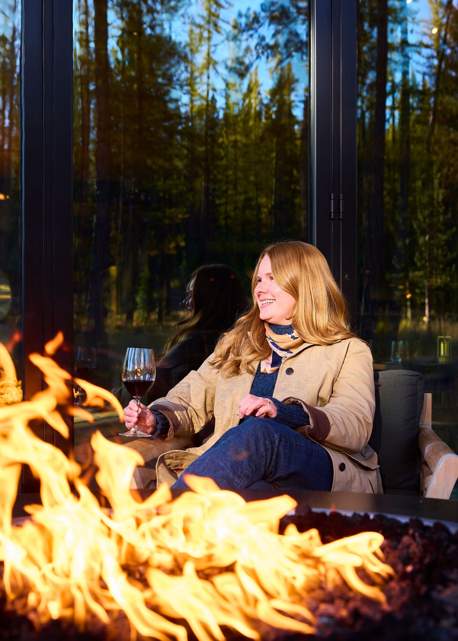 Woman enjoying wine by a fire pit outdoors with forest reflections in glass windows behind her.