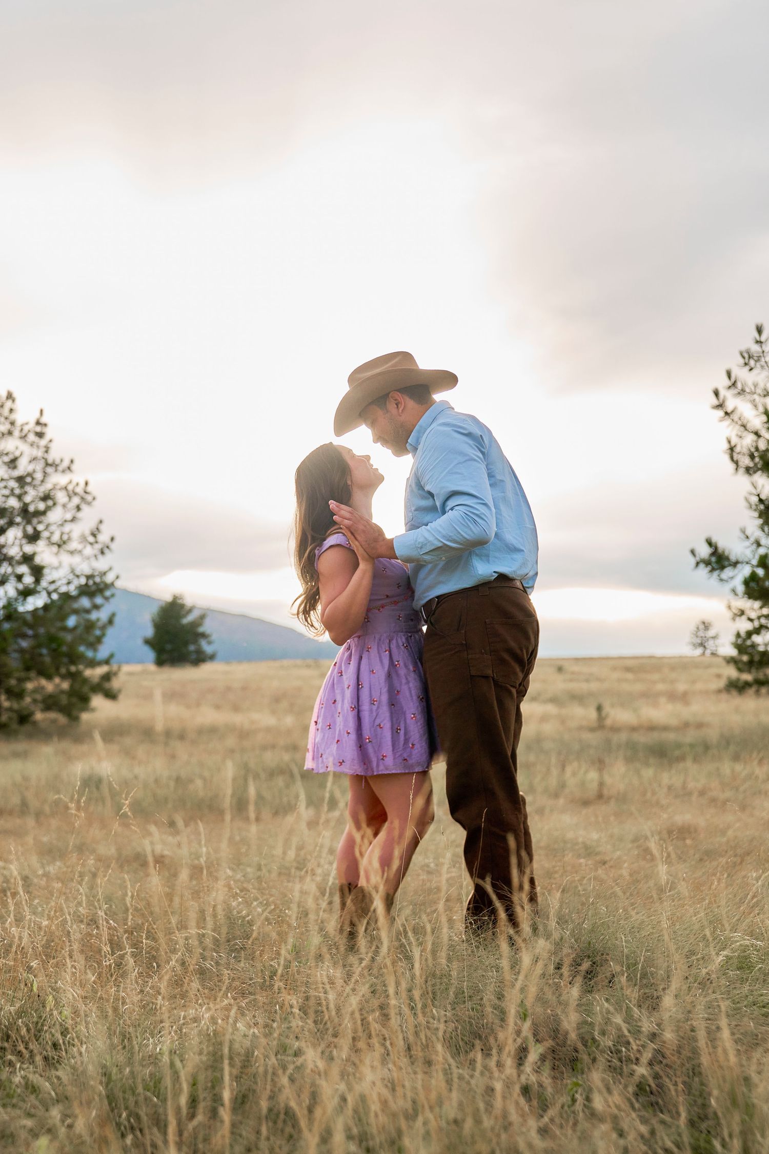 Couple slow dancing in a golden meadow at sunset, man in cowboy hat and blue shirt embracing woman in purple dress.