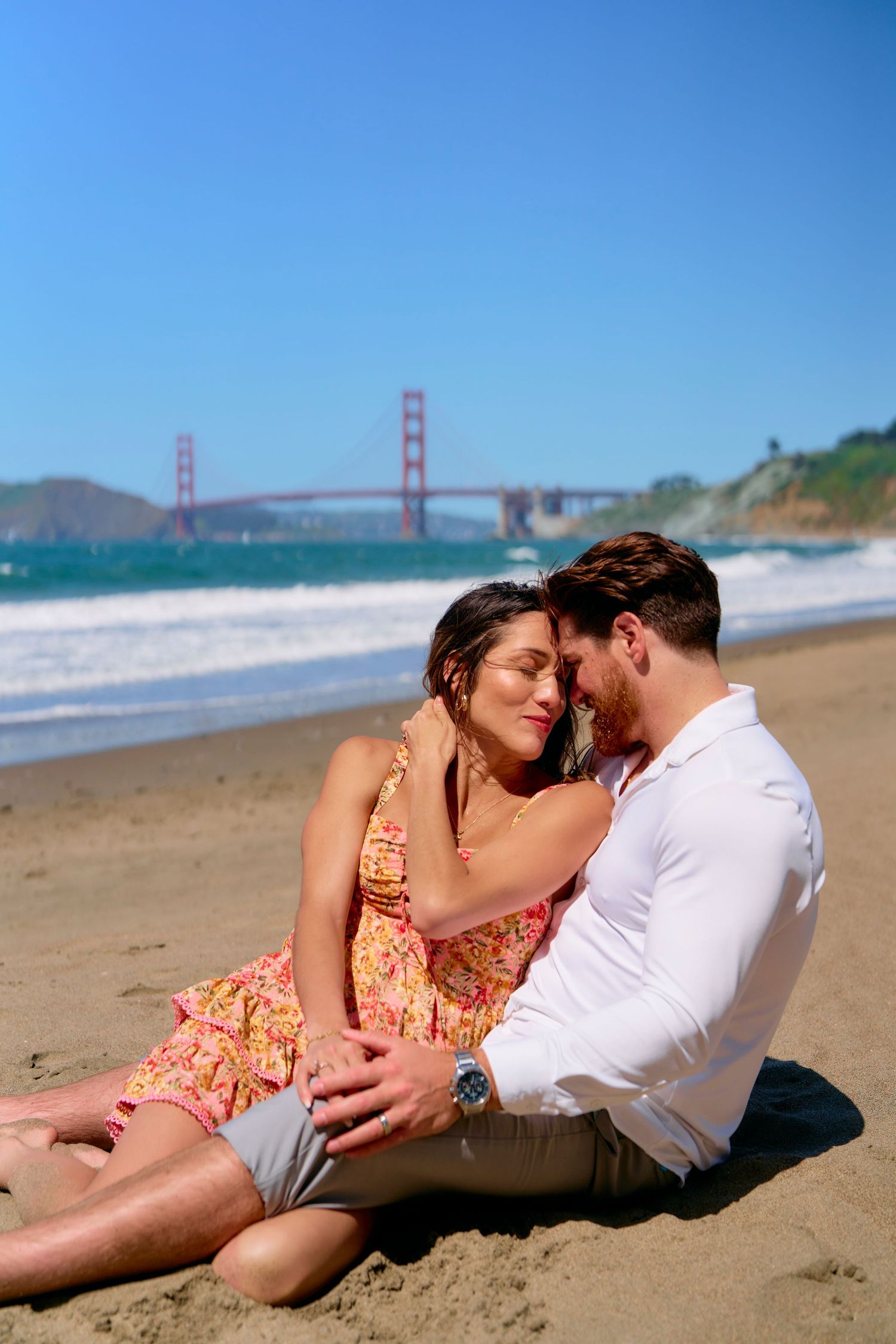 Romantic couple sitting close on Baker Beach with Golden Gate Bridge in sunny background.