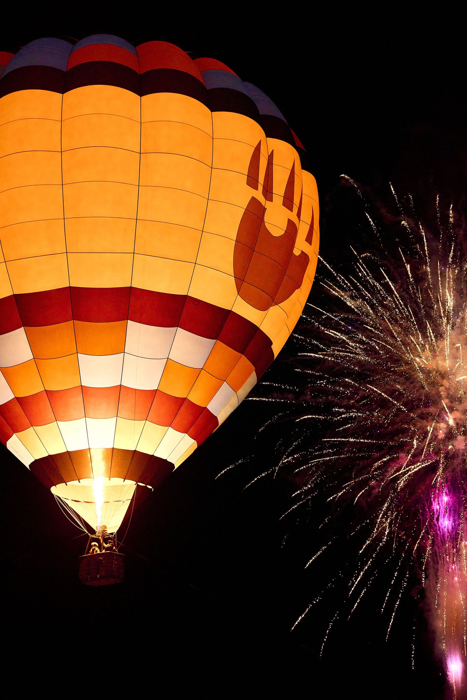 Glowing orange hot air balloon illuminated against night sky with colorful fireworks display.