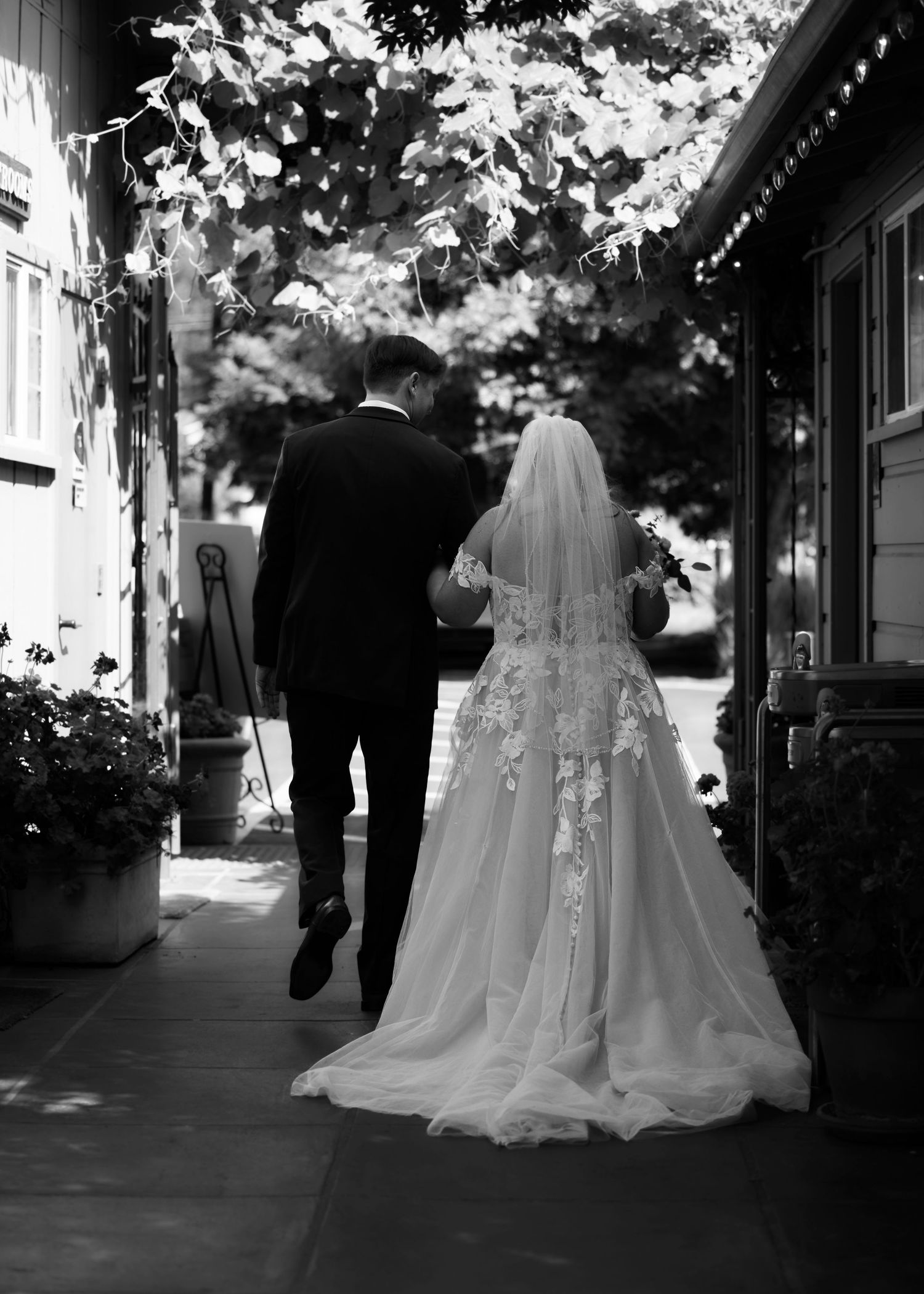 Black and white photo of bride and groom walking hand in hand, bride wearing lace wedding gown with veil.