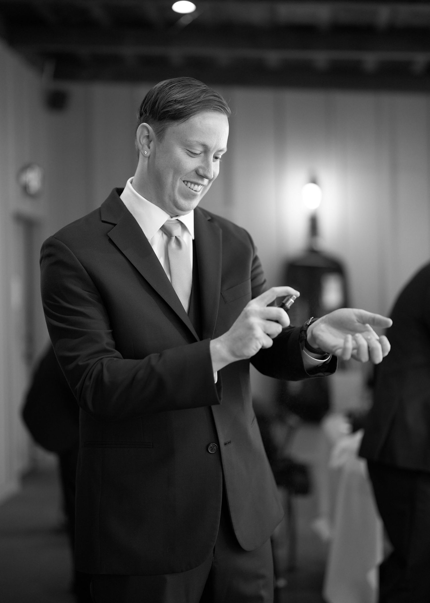 Smiling groom in black suit examines wedding ring in black and white indoor photo.