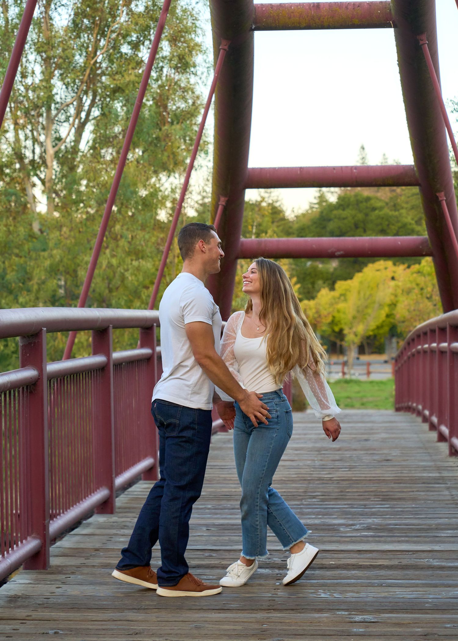 Engaged couple holding hands and smiling on a red wooden bridge in a park.