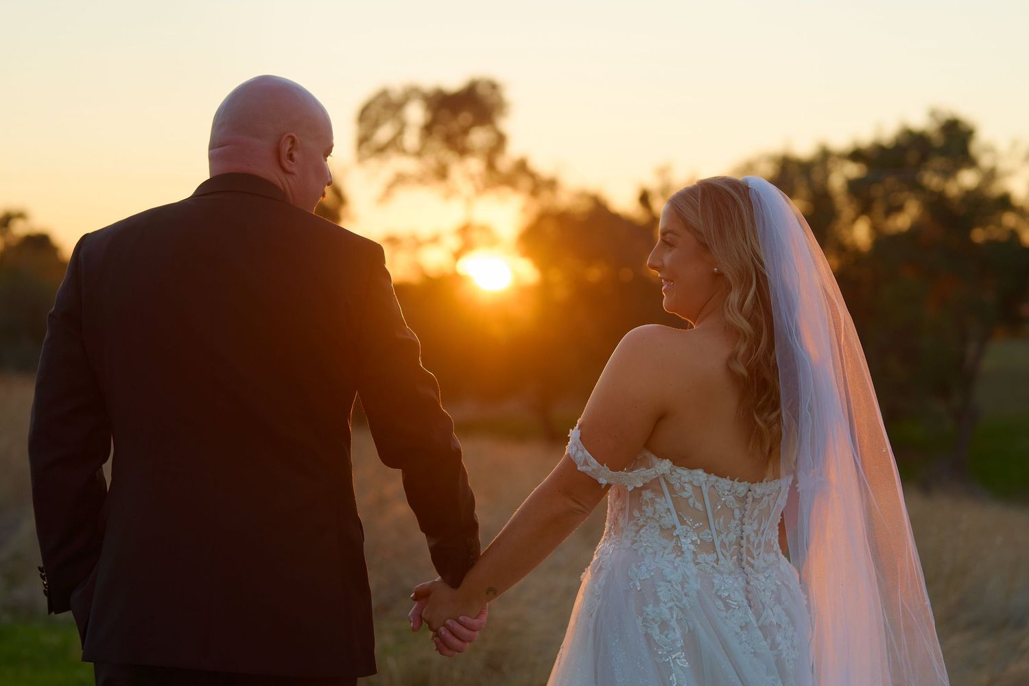 Bride and groom walking hand in hand at sunset, bride in lace gown with veil glancing back warmly.