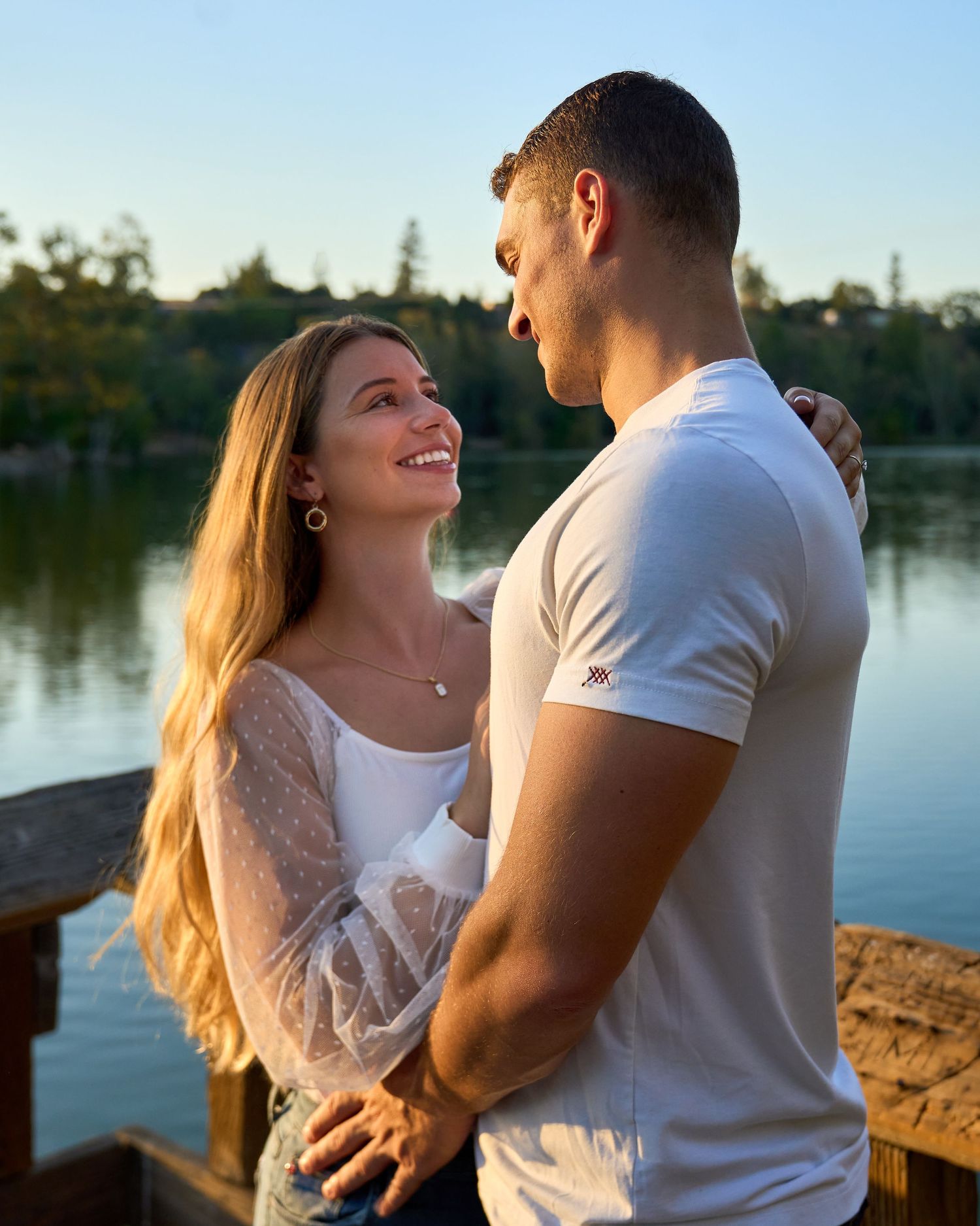 Smiling couple embracing on wooden dock at golden hour with lake and trees behind them.