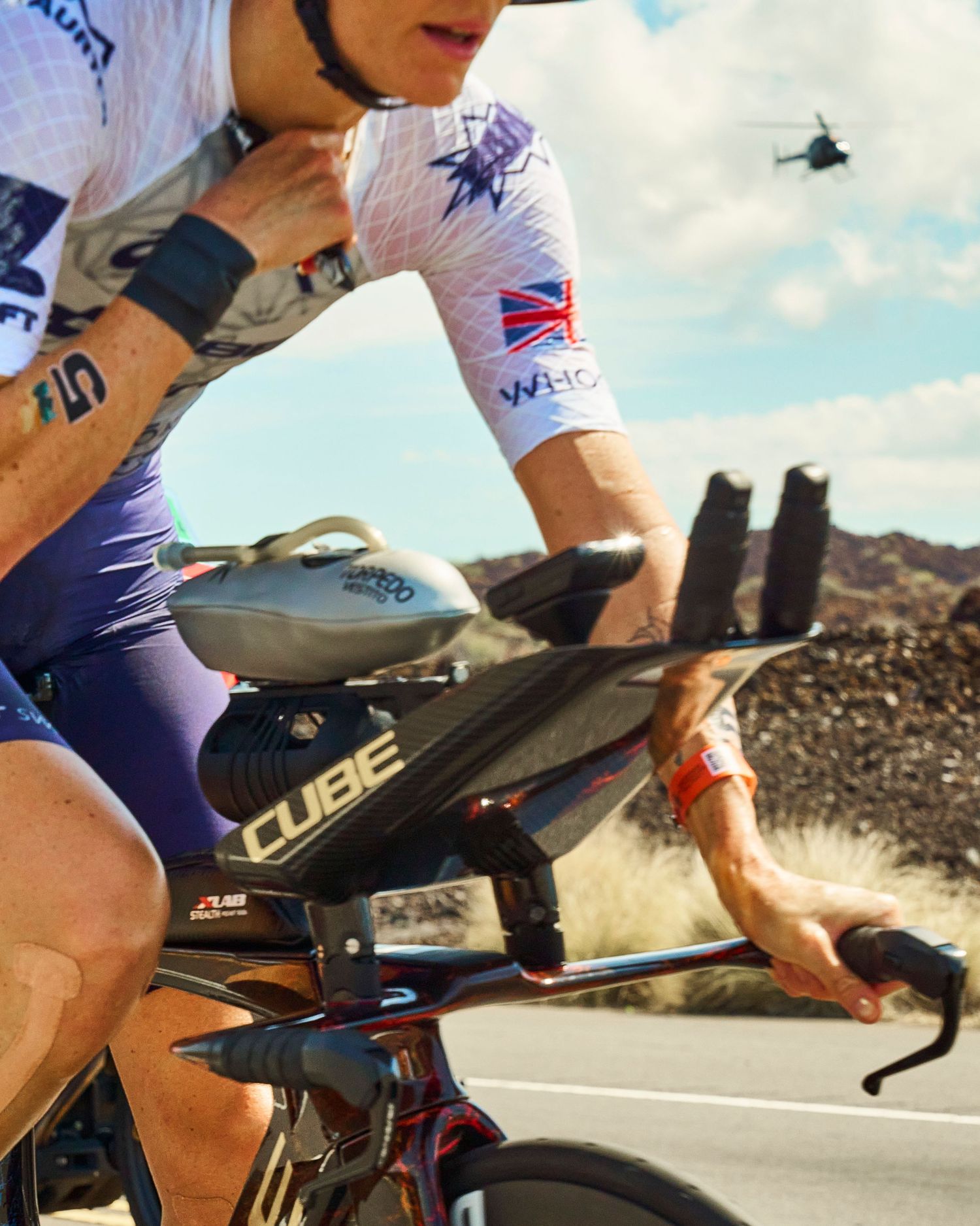 Triathlete in British flag jersey rides a Cube bike in arid landscape with helicopter overhead during a race.