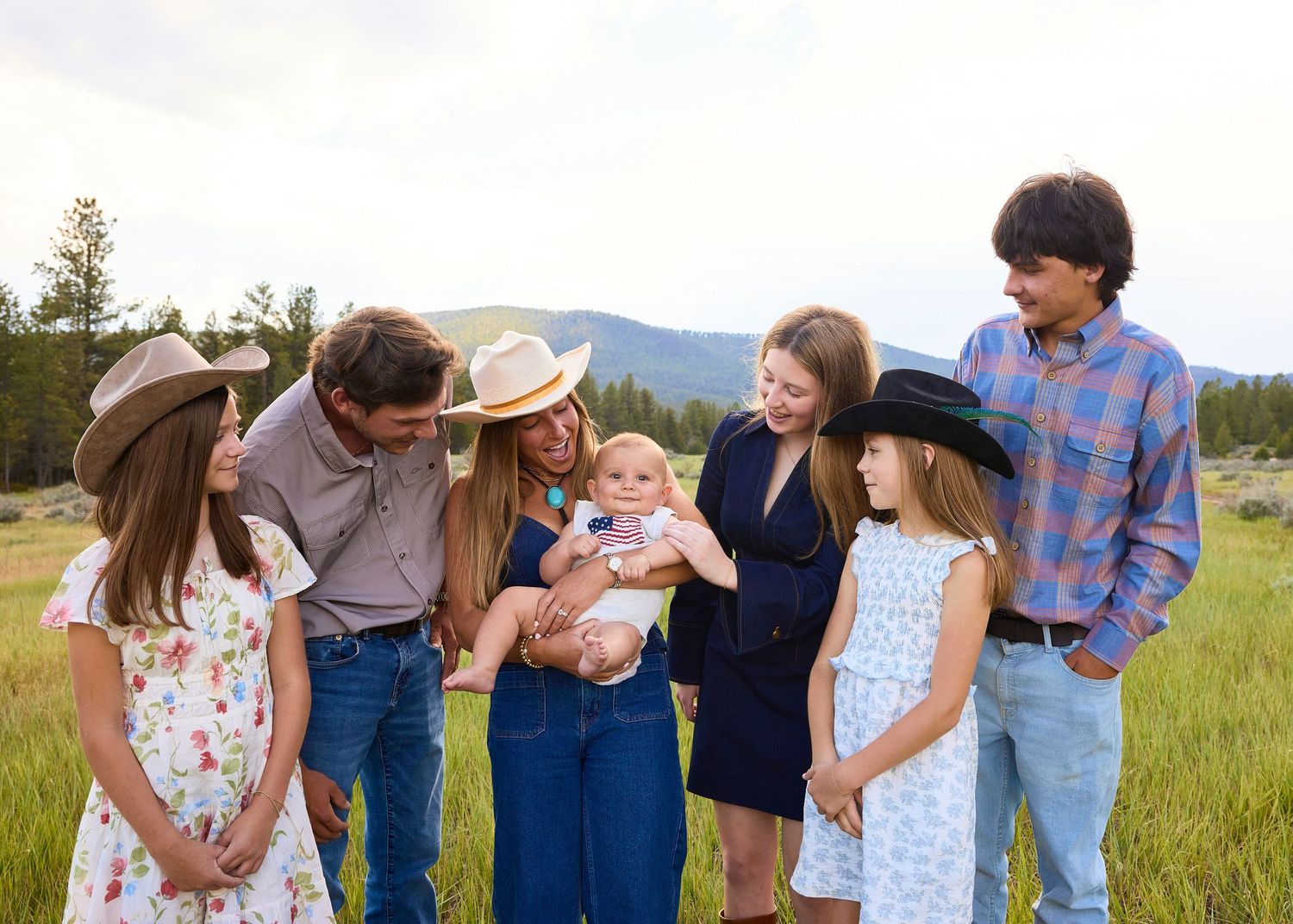 Family group gathered closely in a meadow admiring a smiling baby, with mountain backdrop.