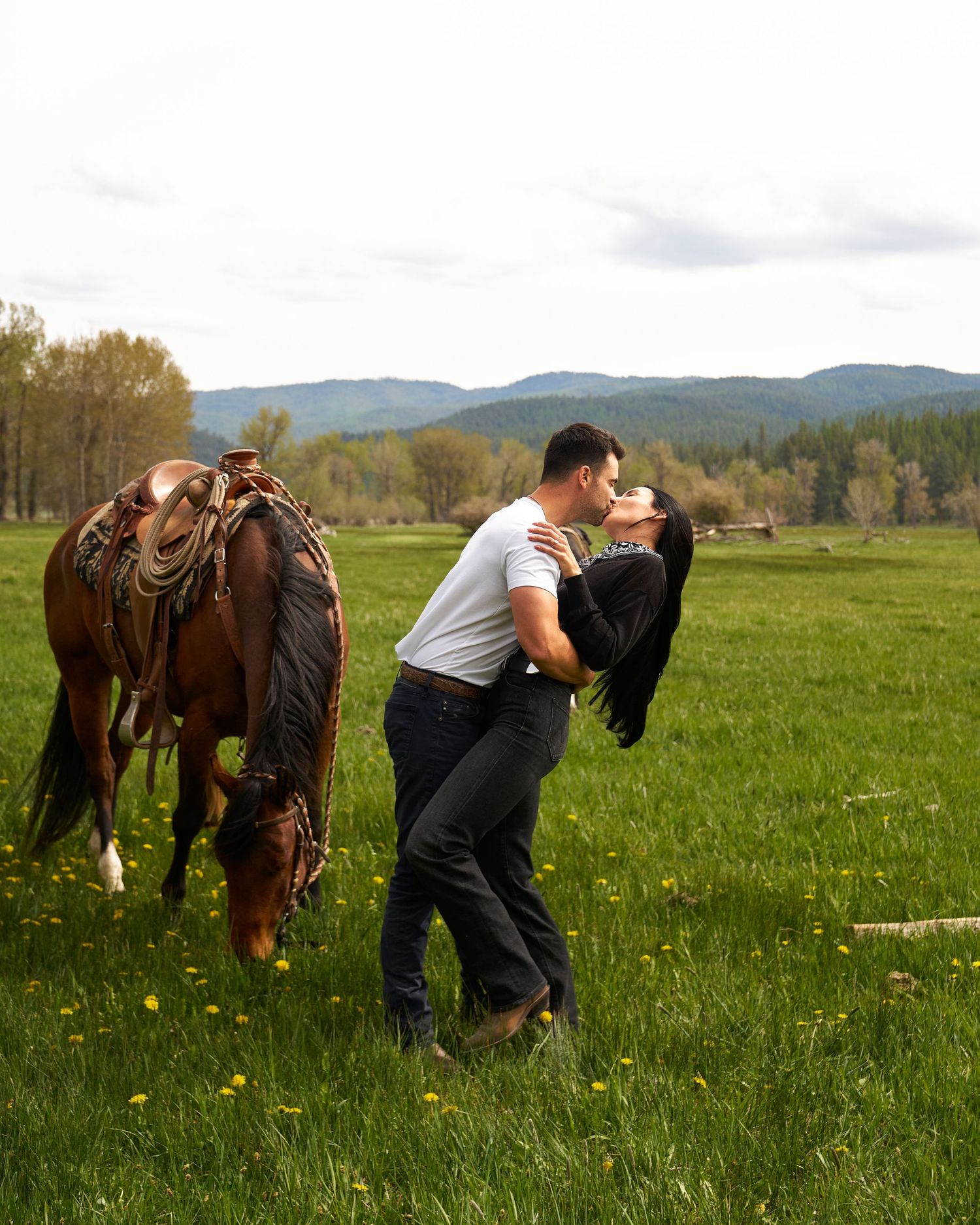 Couple sharing passionate kiss in meadow with grazing horse.